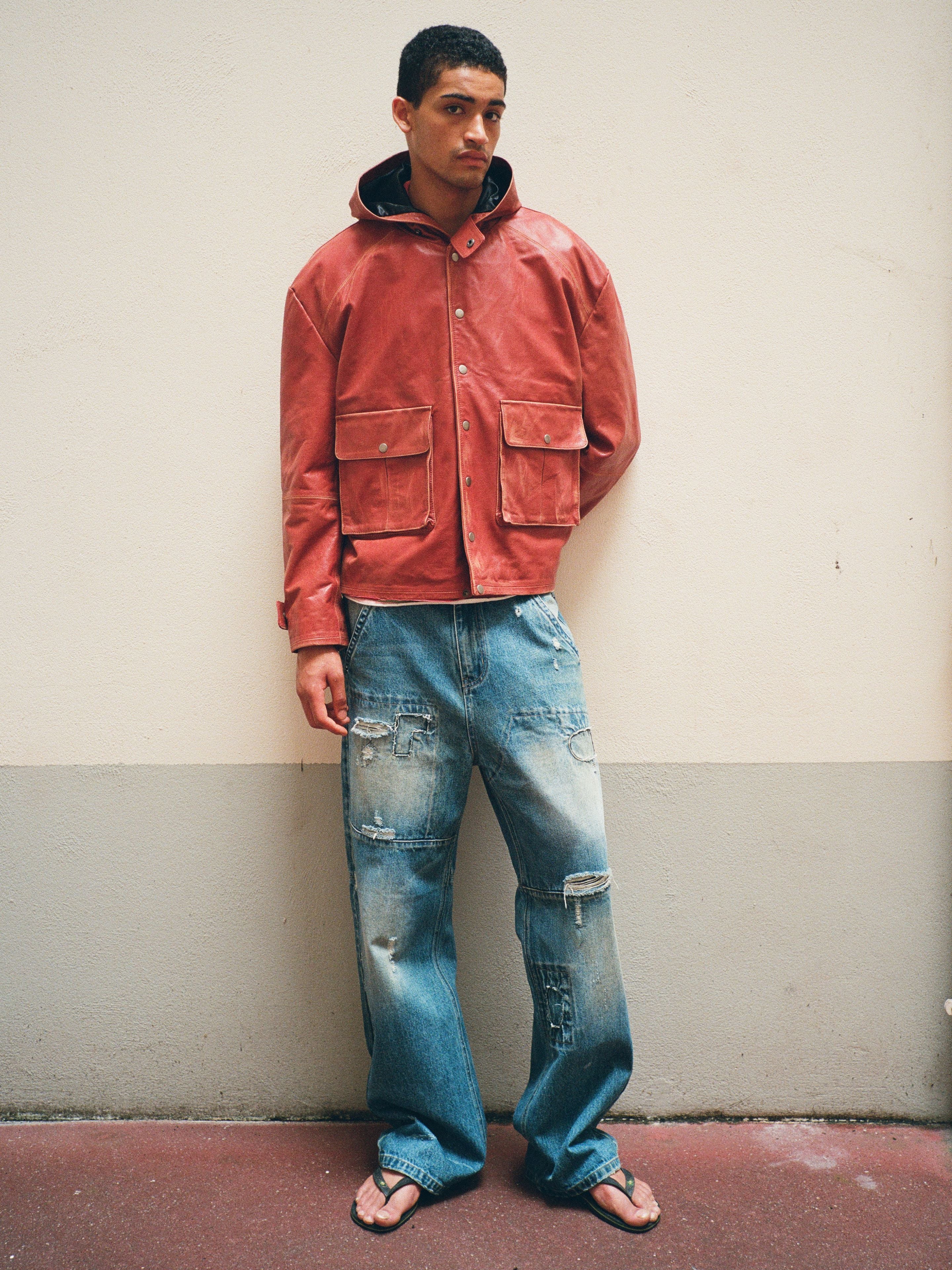 A man stands against a wall wearing a red hooded jacket, FOUND Painter Repaired Jeans, and flip-flops.