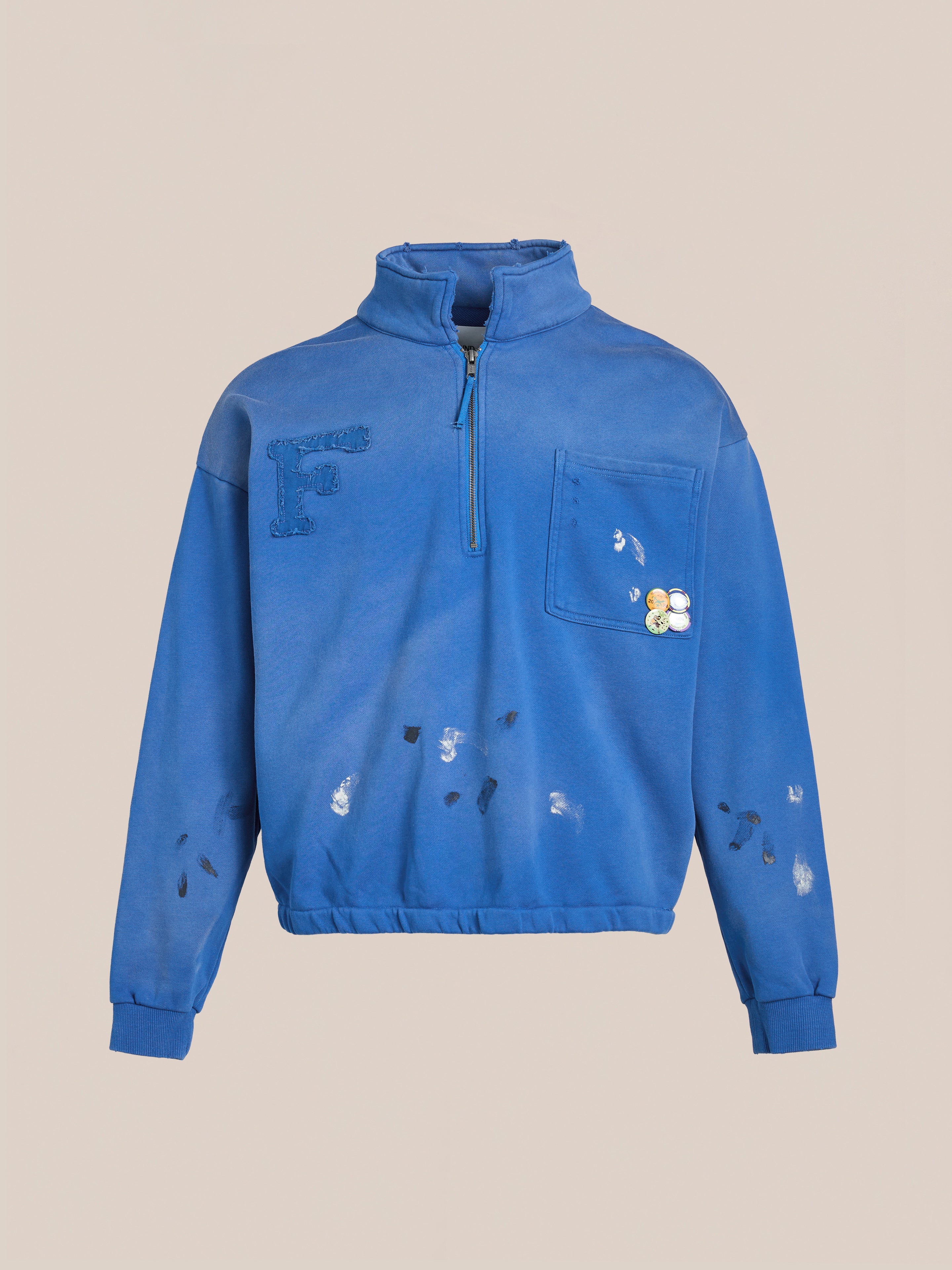 The FOUND Painters Pullover Sweatshirt is a unisex blue quarter-zip with a chest pocket, decorative patches, and workwear style. It features a large "F" among scattered designs on a plain backdrop.