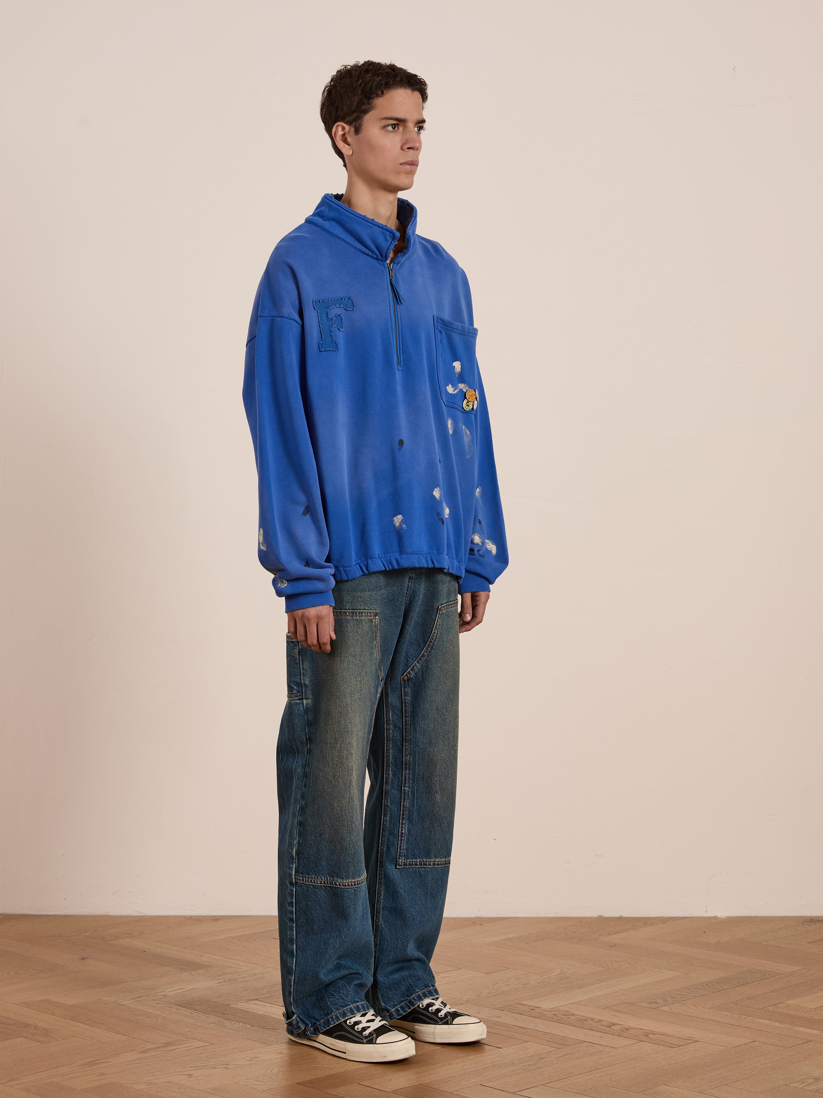 Wearing the Painters Pullover Sweatshirt from FOUND, teamed with wide jeans, a person in a blue jacket decorated with patches stands on a wooden floor, slightly facing right—a nod to unisex workwear and casual elegance.