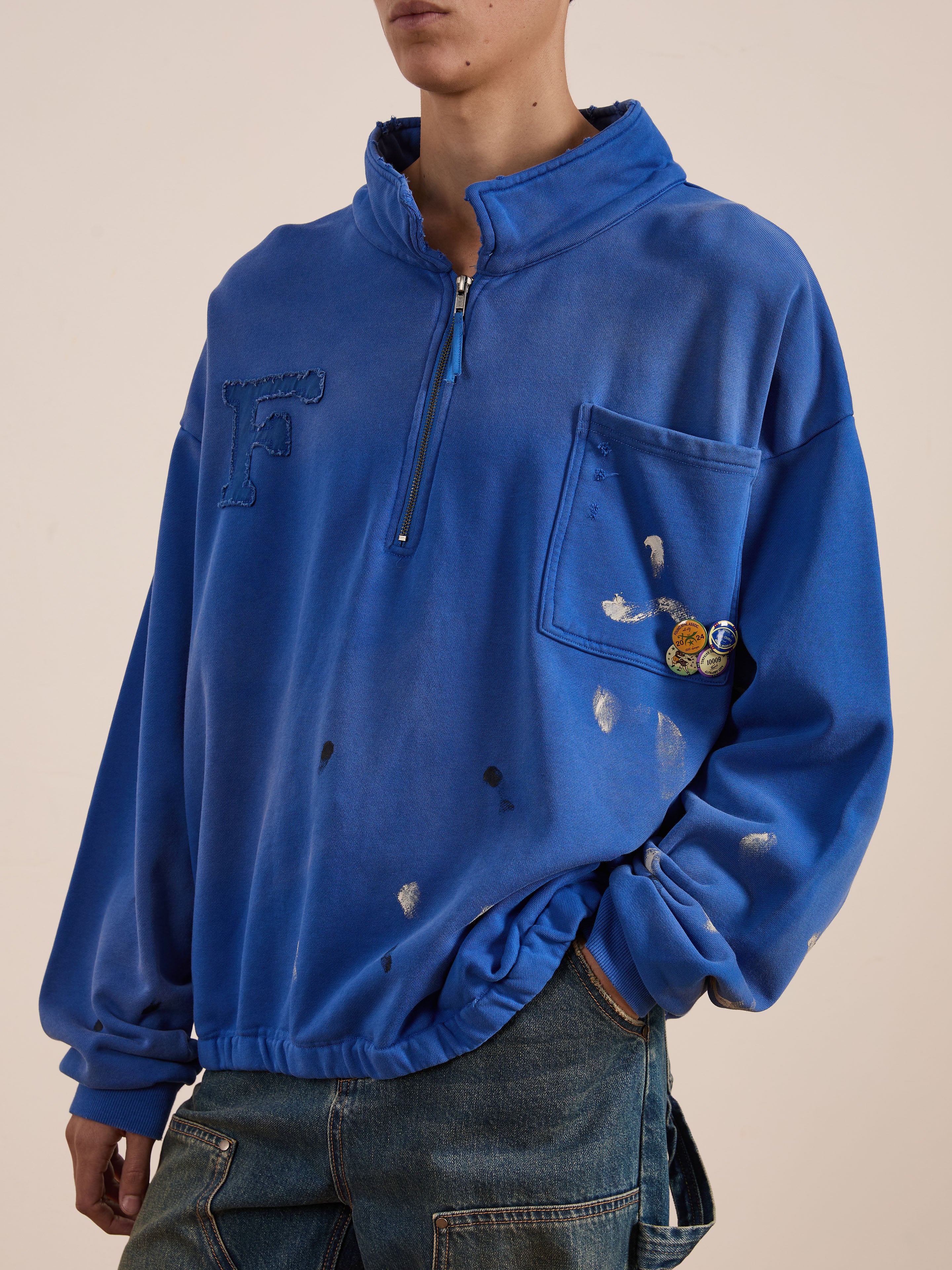 Someone is wearing the Painters Pullover Sweatshirt by FOUND. This unisex blue zip-up features a pocket and buttons, enhanced with paint splatters for an artistic, workwear-inspired look.