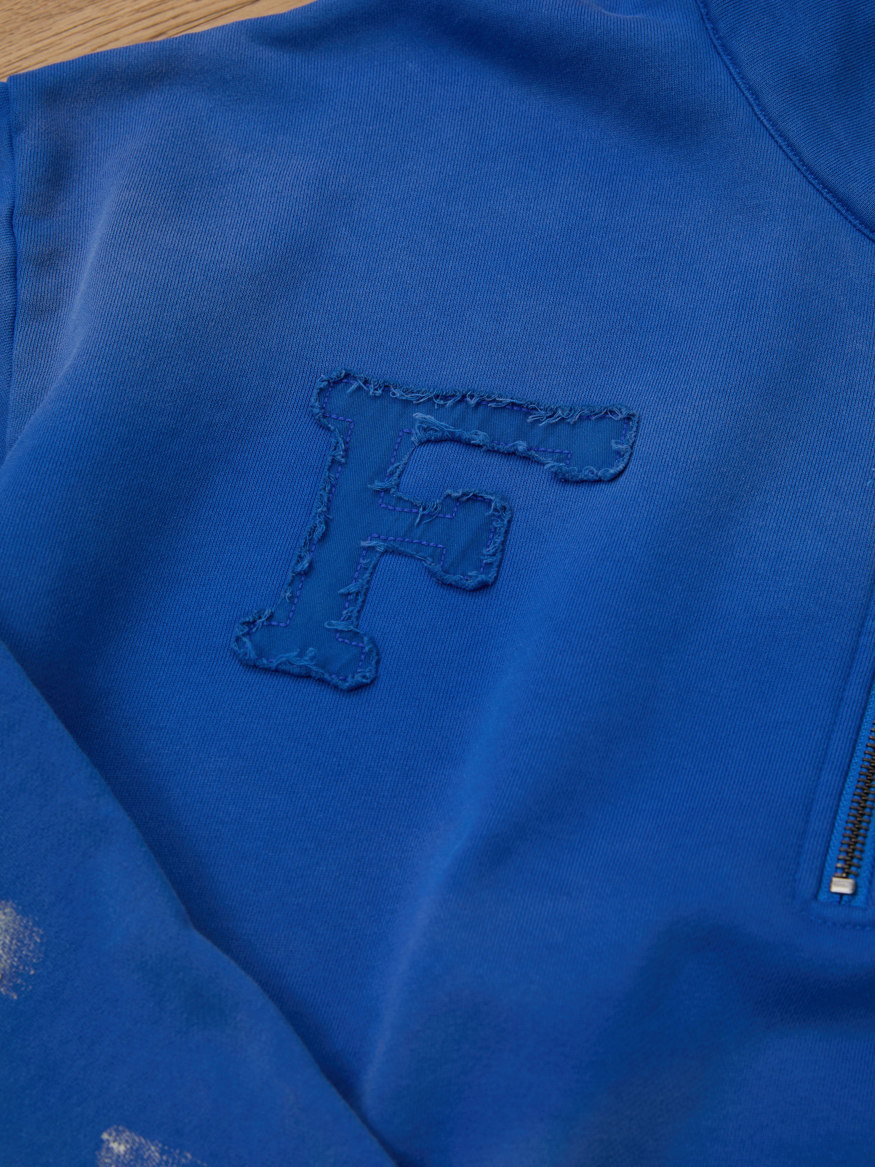 Close-up of the Painters Pullover Sweatshirt by FOUND, showcasing a blue design with an "F" stitched on the chest and a partial zipper on the right. This unisex workwear piece blends comfort and style seamlessly.