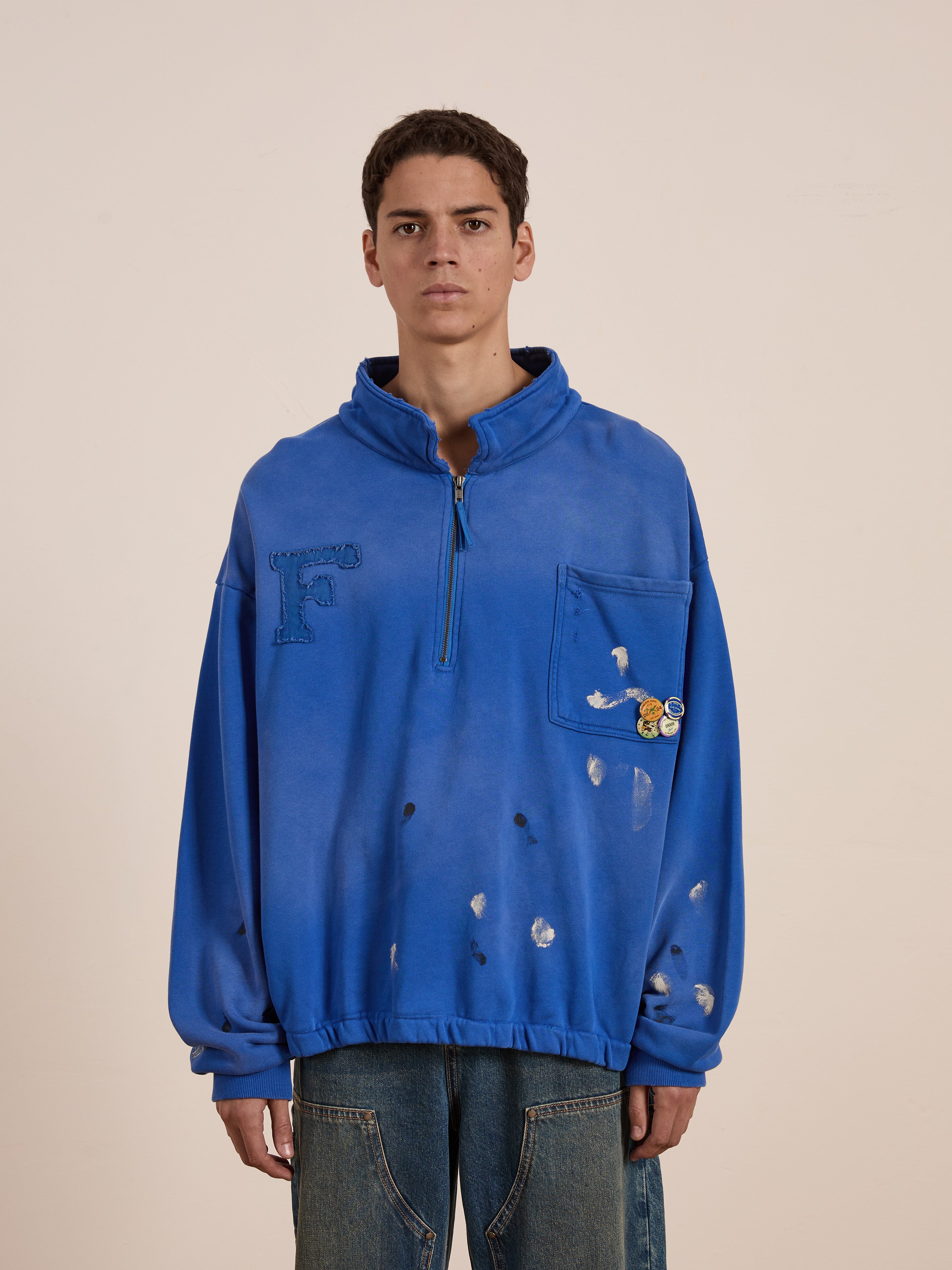 A person is wearing the Painters Pullover Sweatshirt by FOUND, a unisex blue pullover with a front pocket, splatter mark designs, and playful pins. This sweatshirt offers casual comfort with a workwear-inspired style.