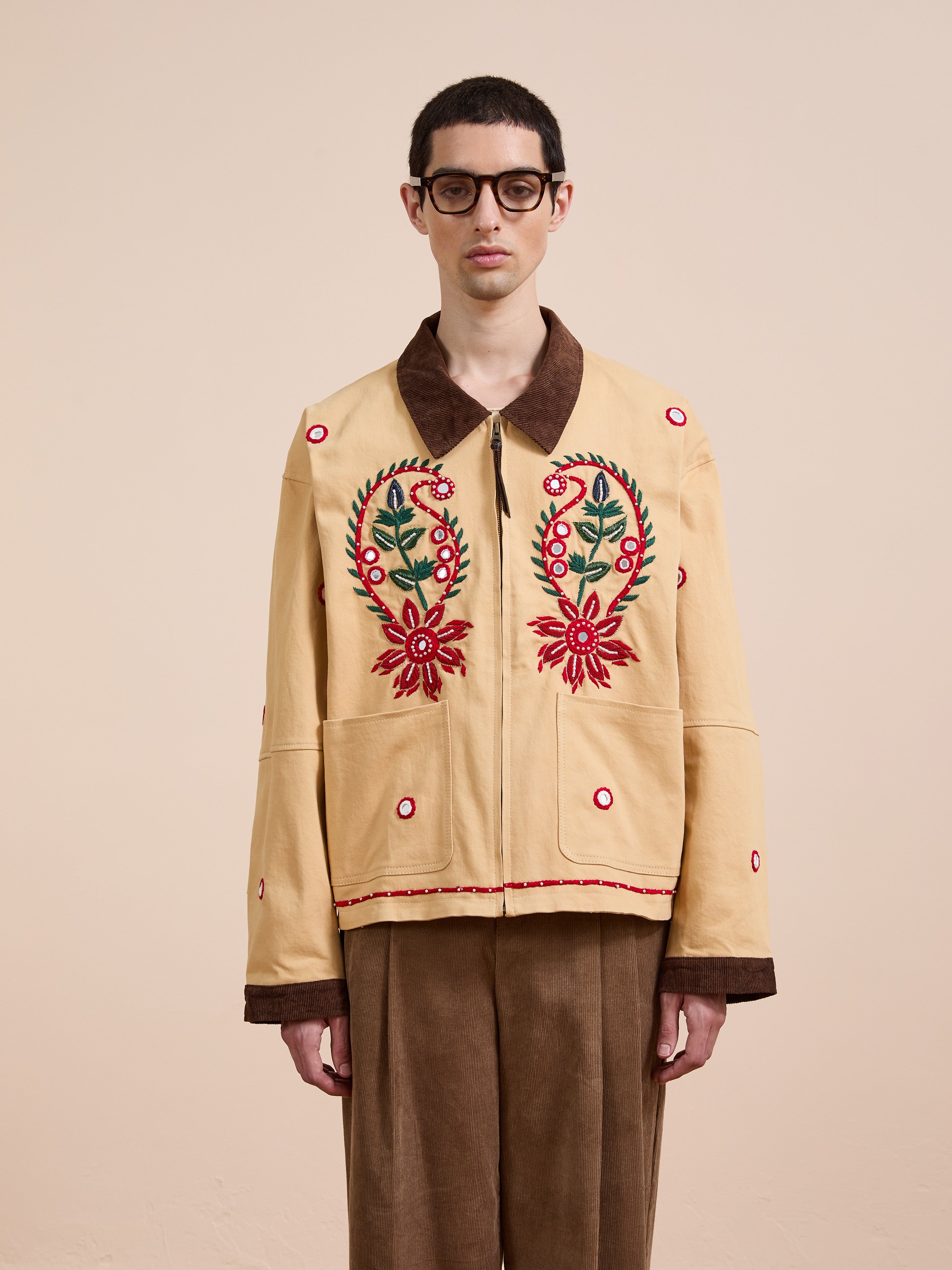 A person wearing glasses and brown trousers stands facing forward against a beige background, showcasing Found's Paisley Motif Hand Embroidered Work Jacket with red floral embroidery and artisan craftsmanship.