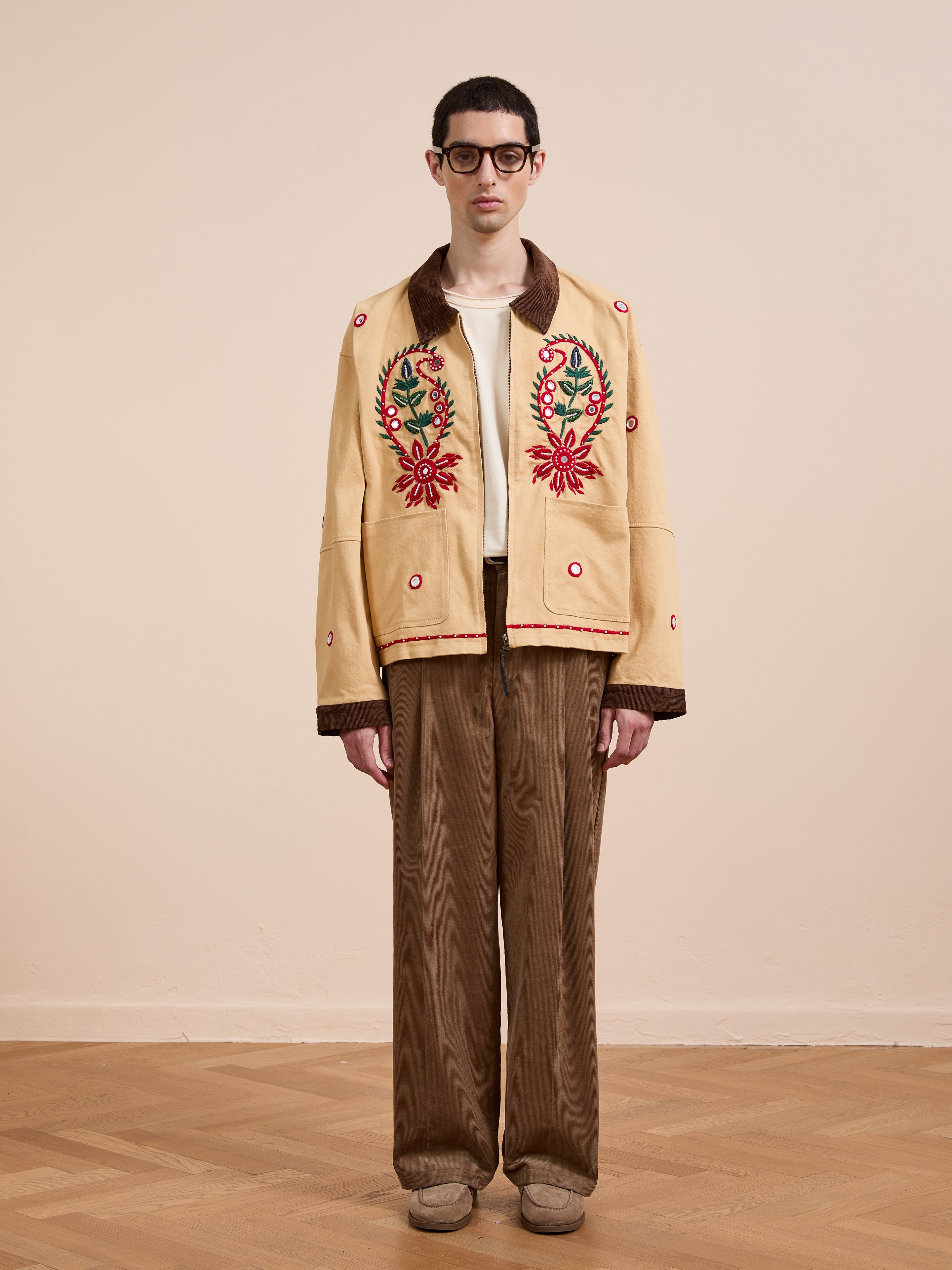 A man in glasses stands against a plain wall, wearing Found’s Paisley Motif Hand Embroidered Work Jacket over a white shirt, paired with brown corduroy pants and beige shoes.