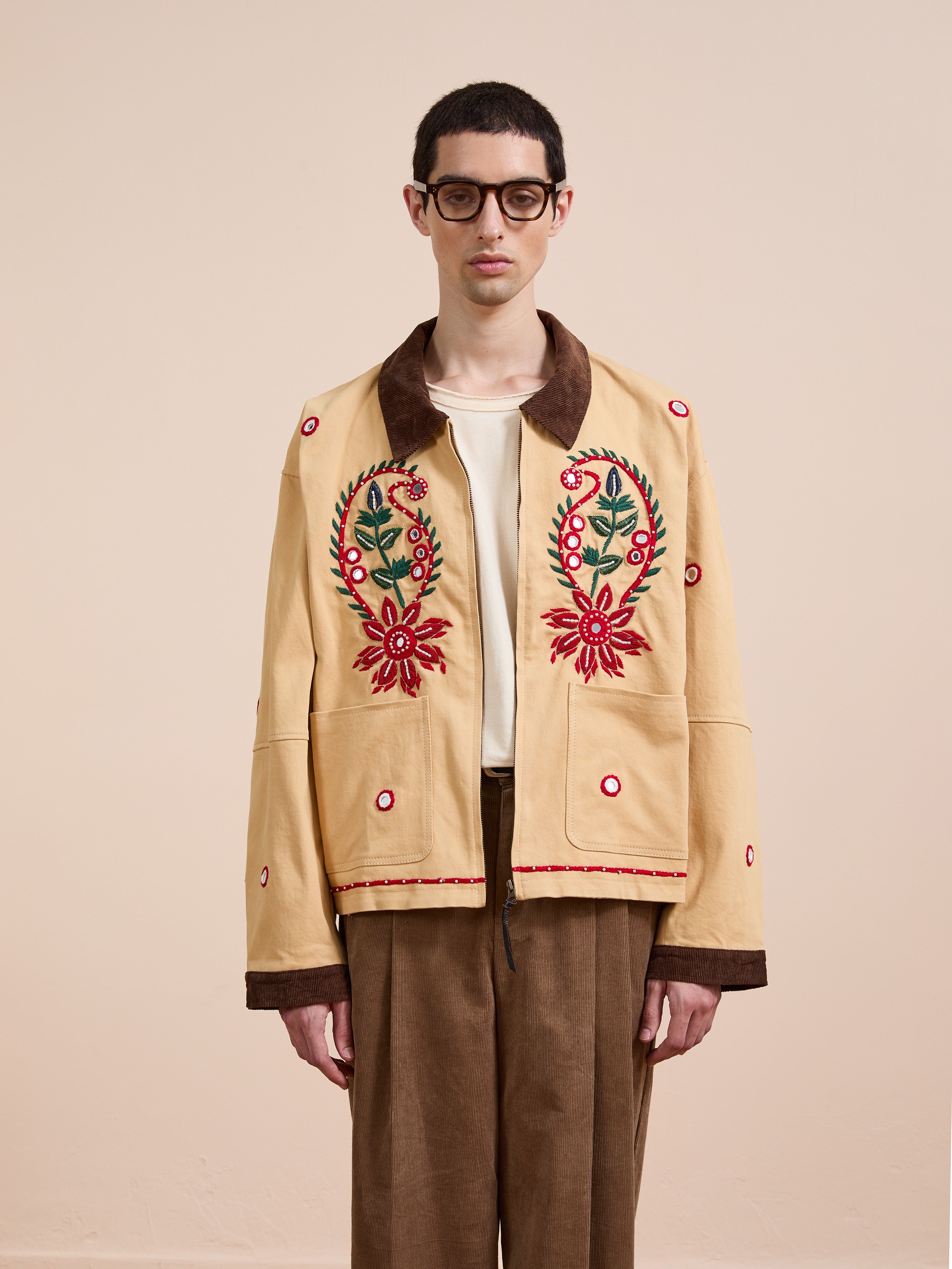A person wearing glasses and Found's Paisley Motif Hand Embroidered Work Jacket in beige with red floral embroidery and brown trim, paired with brown pants, stands against a plain background, highlighting artisan craftsmanship.