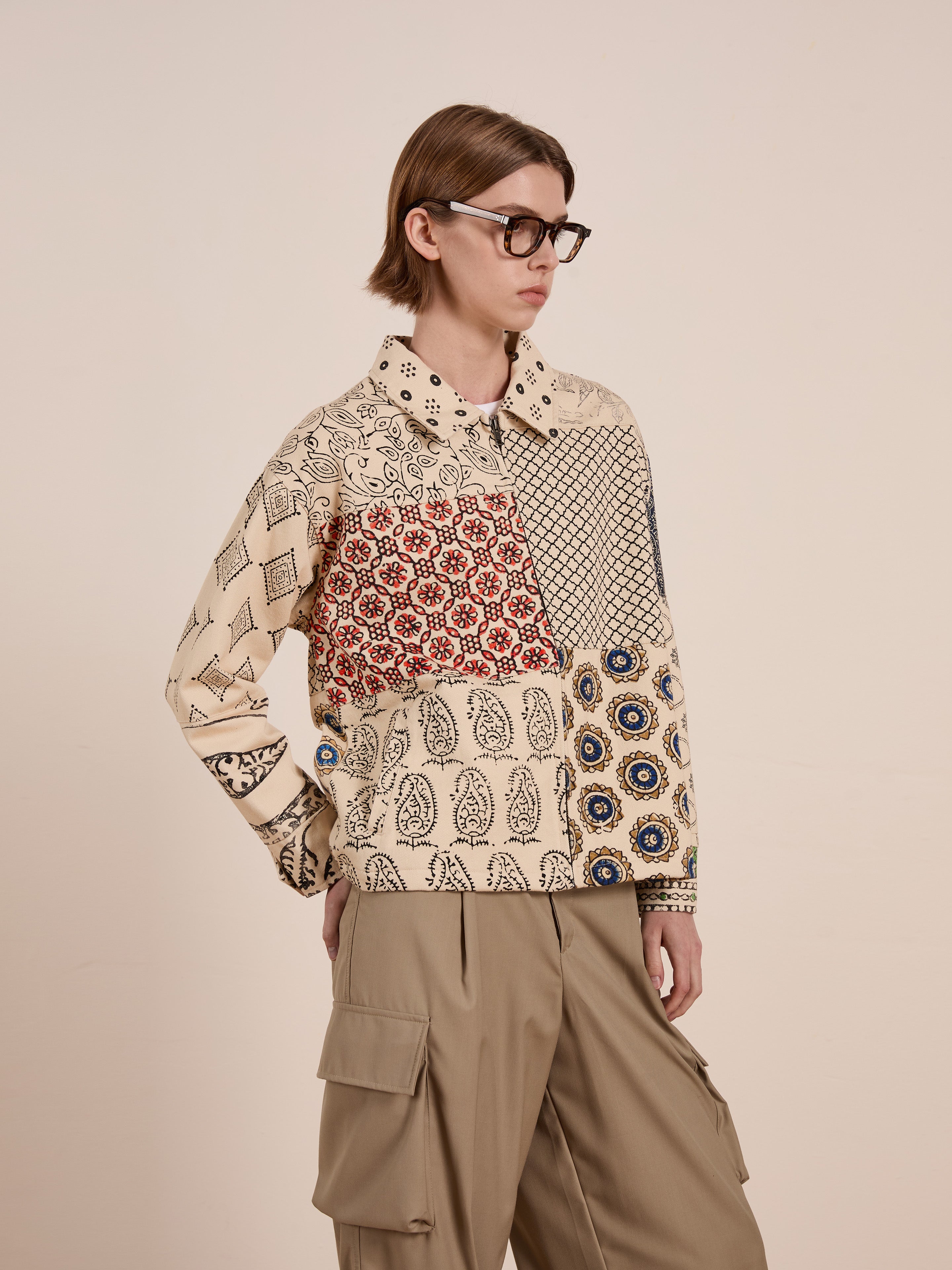 A person wearing glasses, a FOUND Paisley Mosaic Work Jacket, and beige cargo pants stands with one hand in their pocket against a plain background. This unisex jacket adds a versatile flair to the ensemble.