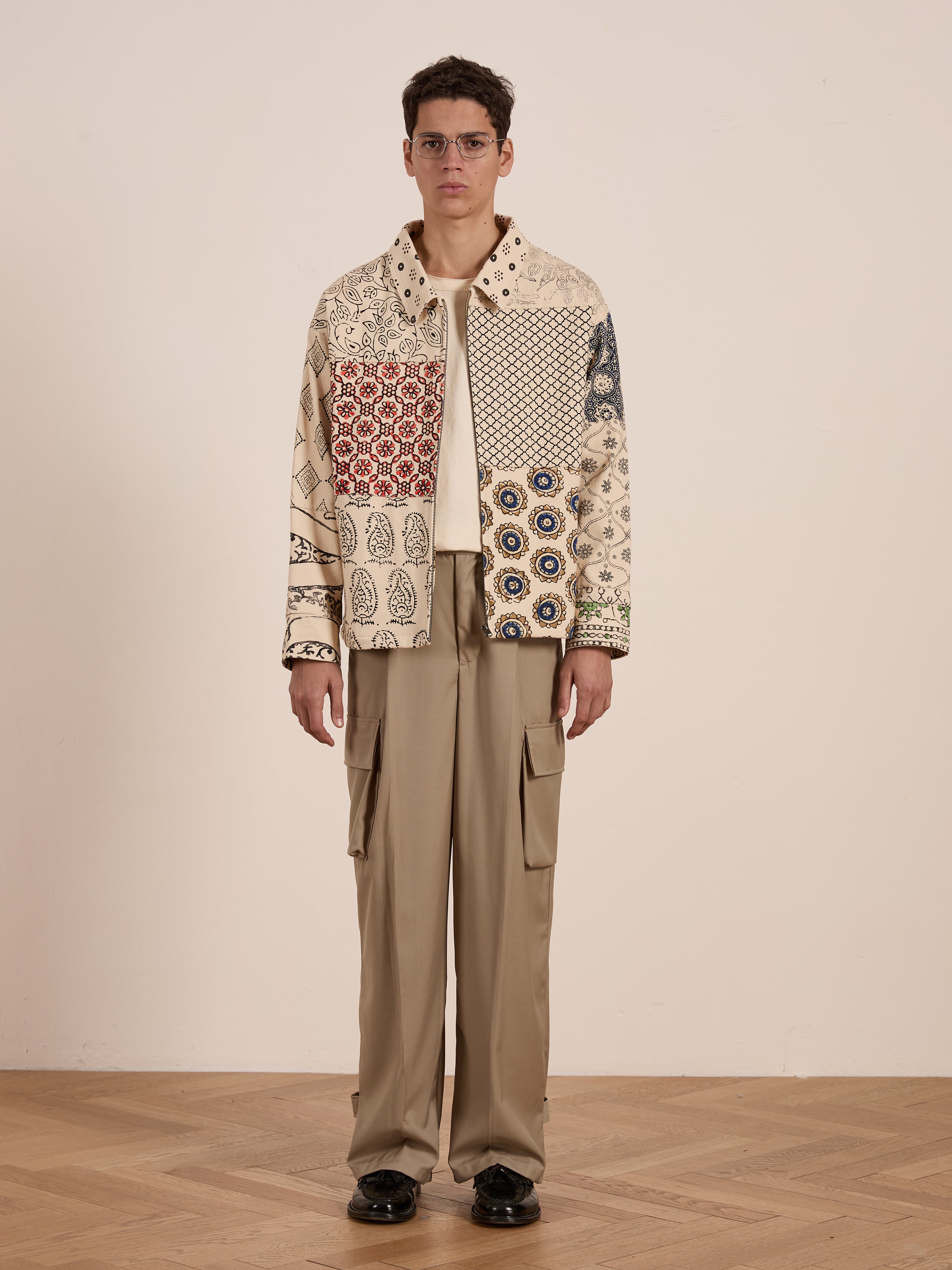 An individual indoors sports a FOUND Paisley Mosaic Work Jacket paired with beige cargo pants and black shoes, set against a plain backdrop.