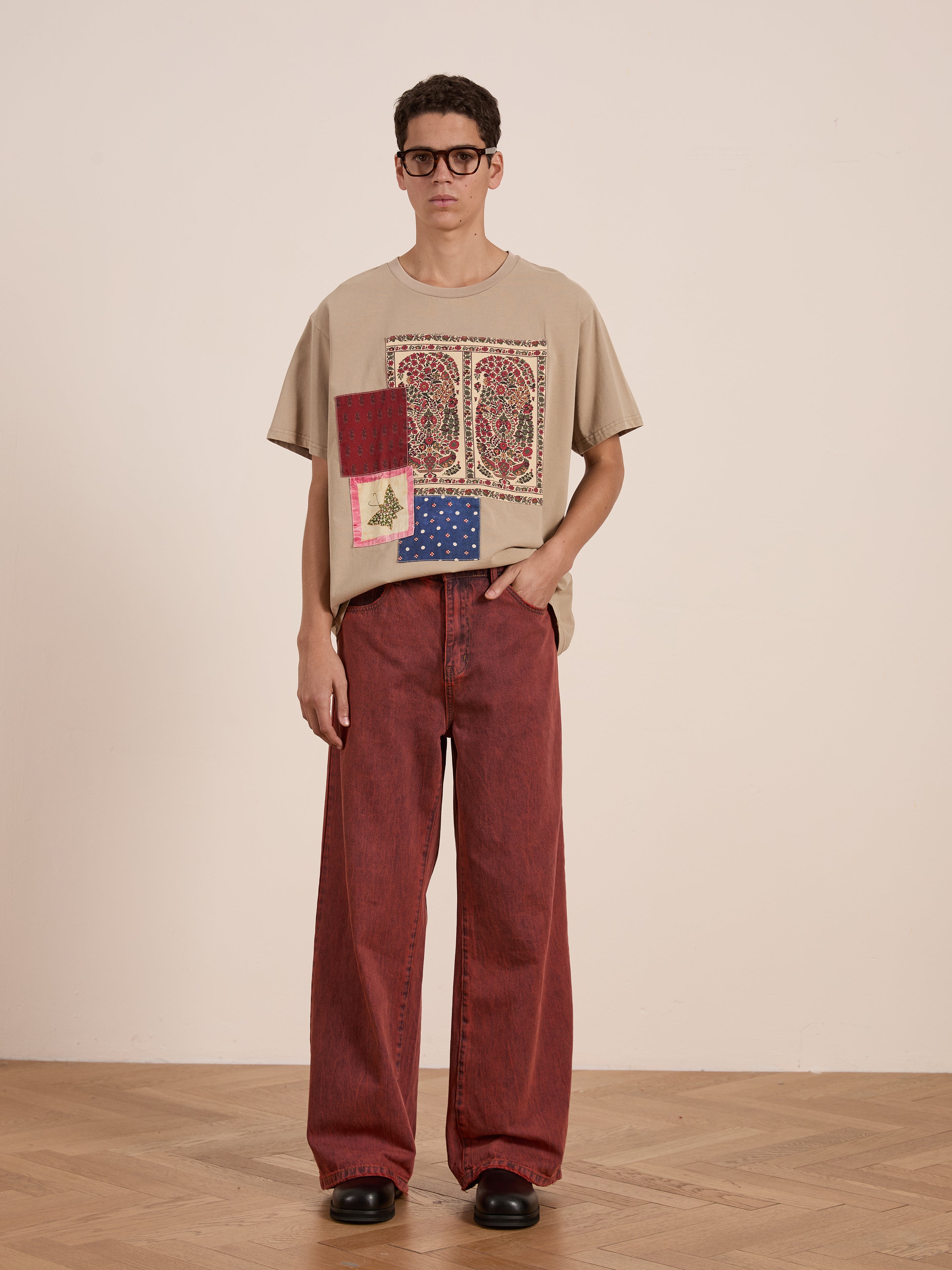 A person wearing glasses and a beige FOUND Patchwork Tee inspired by South Asian quilts, paired with red wide-leg pants and black shoes, stands on a wooden floor against a plain white wall.