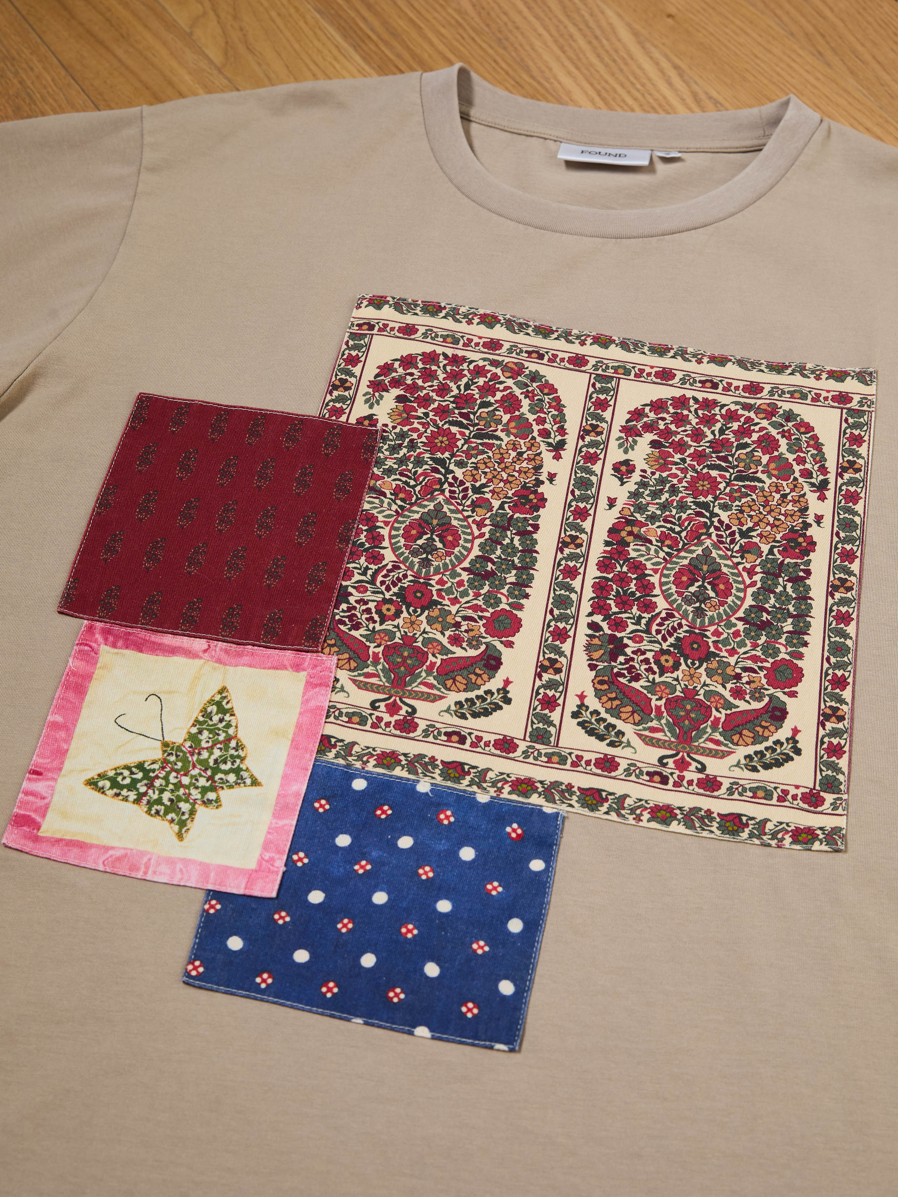 The Patchwork Tee by FOUND is a beige T-shirt featuring a floral pattern inspired by South Asian quilts, displayed with three fabric swatches: floral on red, butterfly on yellow, and polka dots on blue, all set against a wooden surface.