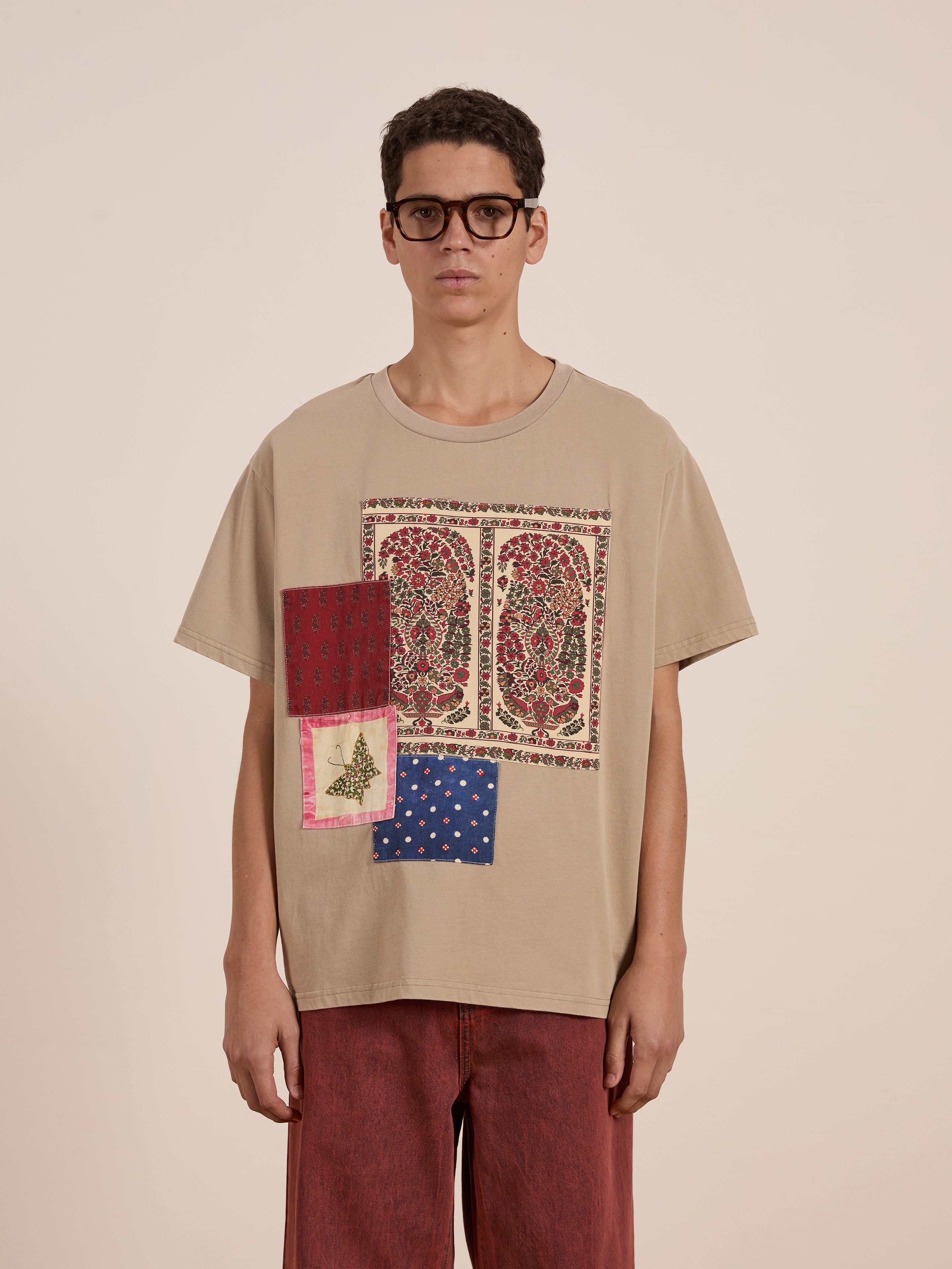 A person wearing glasses and a beige FOUND Patchwork Tee, adorned with vibrant colors as found in South Asian quilts, confidently pairs it with striking red pants against a plain backdrop.