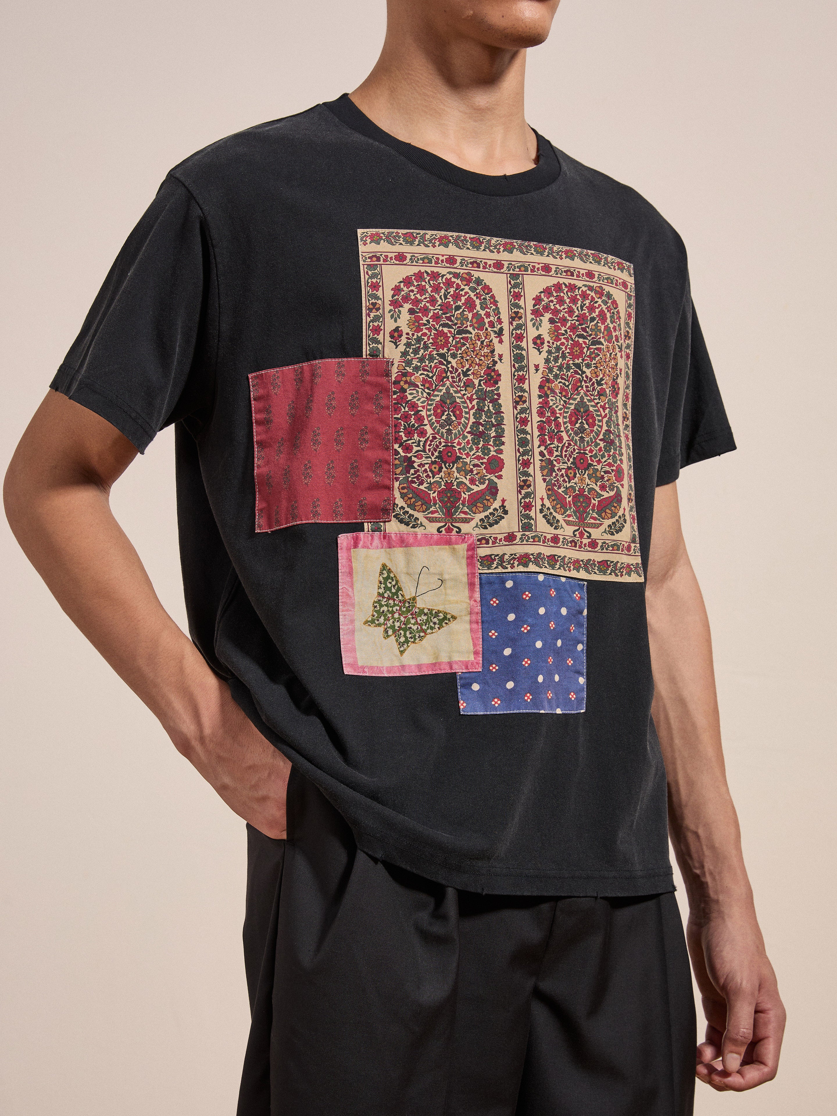 A person wearing the FOUND Patchwork Tee, adorned with decorative patches in various patterns and colors such as red, blue, and multicolored designs, stands with one hand in their pocket.