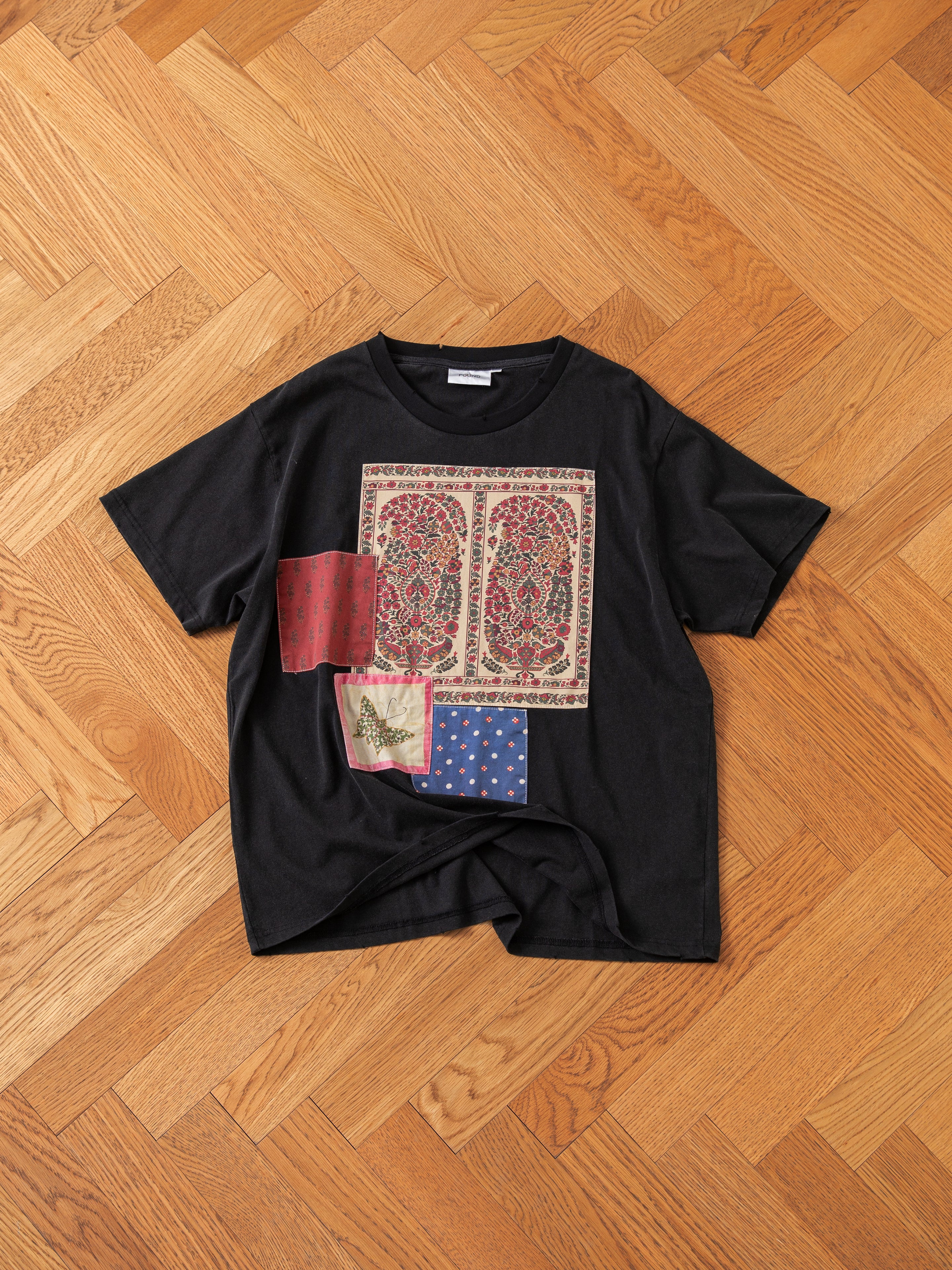 A black short-sleeve Patchwork Tee by FOUND, featuring red, blue, and multicolored patches in a vibrant design, is laid flat on a wooden herringbone floor.