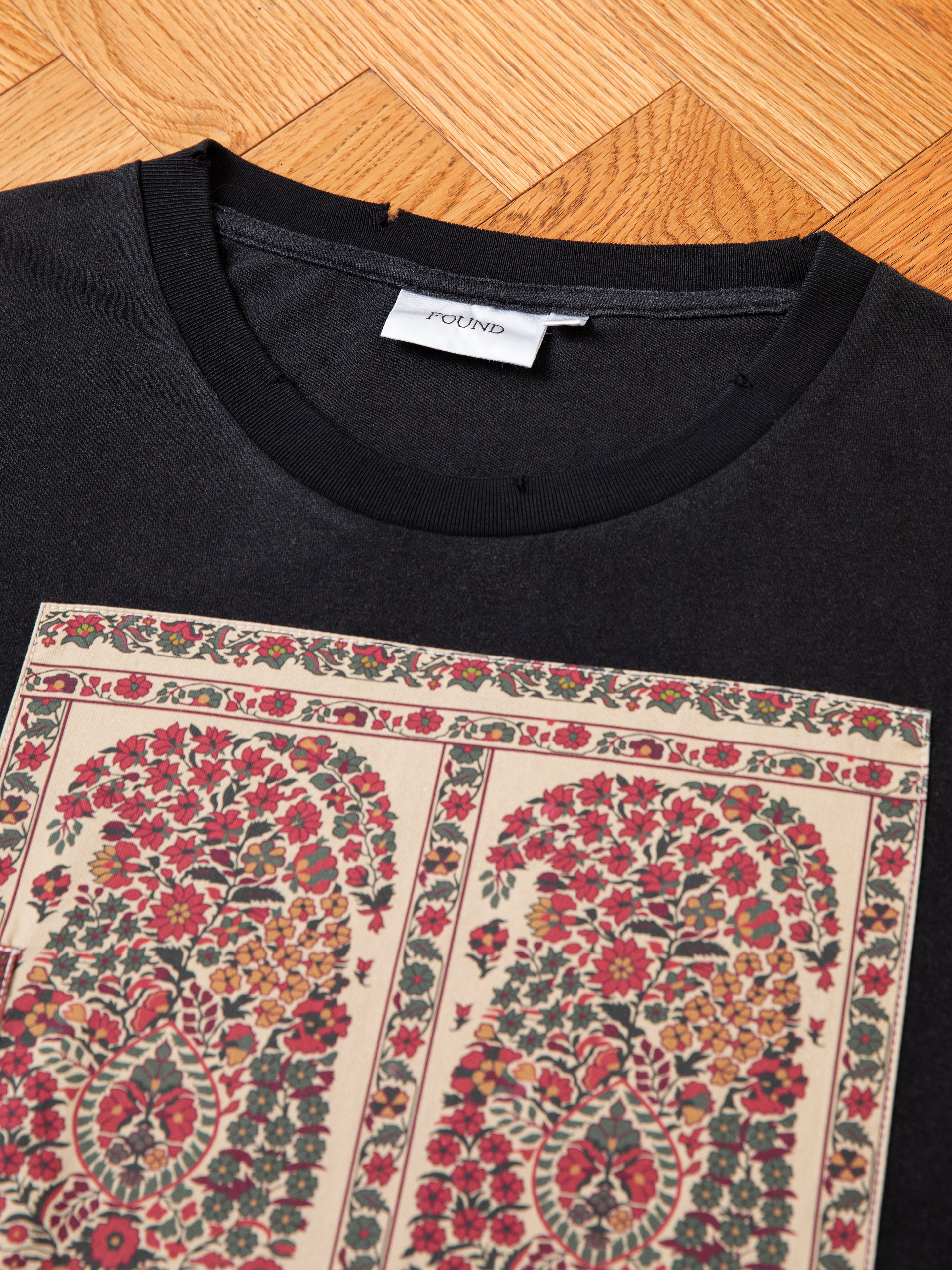 A black Patchwork Tee with a multicolored floral design laid flat on a wooden surface. A white clothing label with the text "FOUND" is sewn inside the collar.