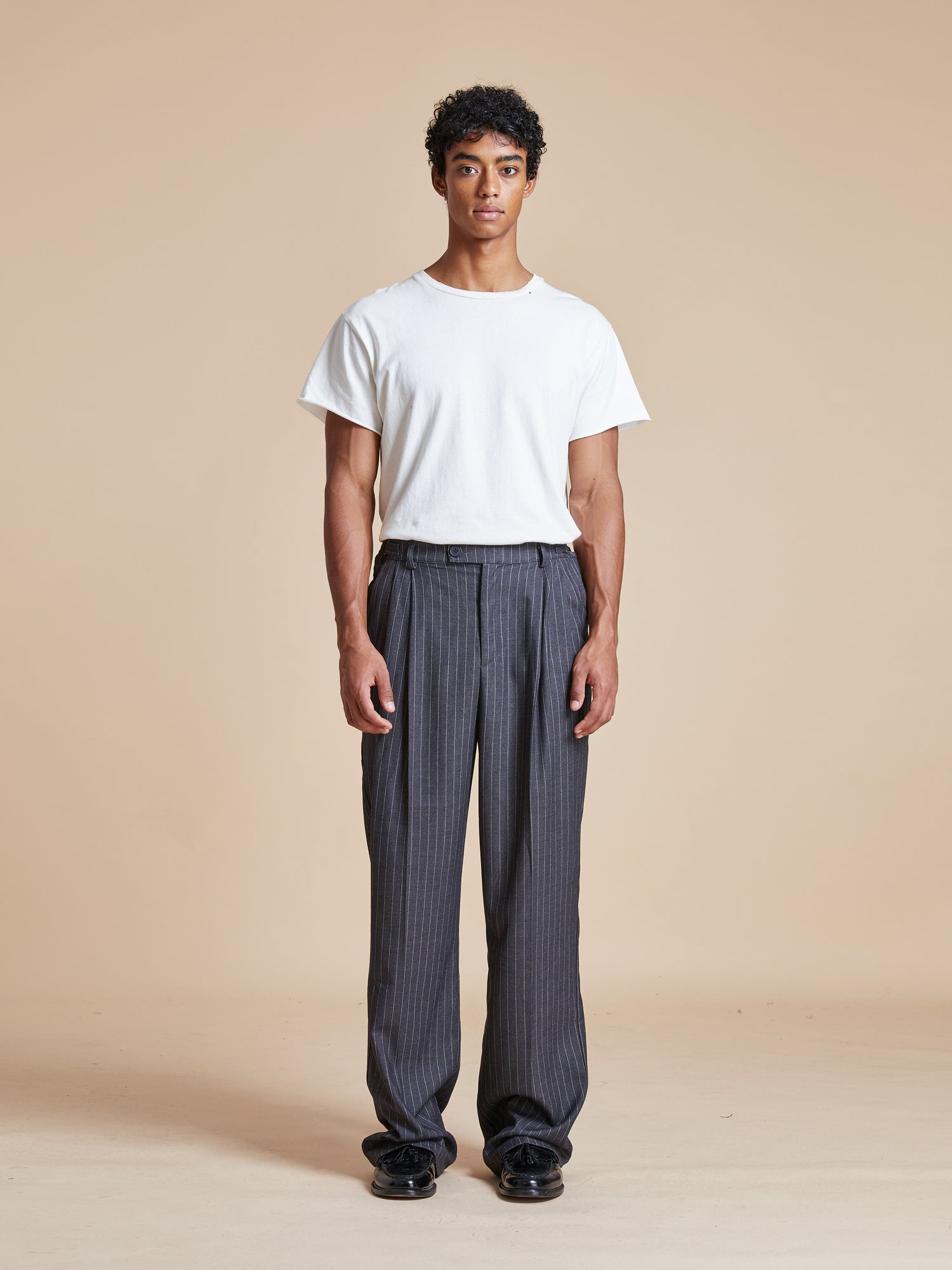 The model is wearing a white t-shirt and grey Found Pinstripe Pleated Trousers.