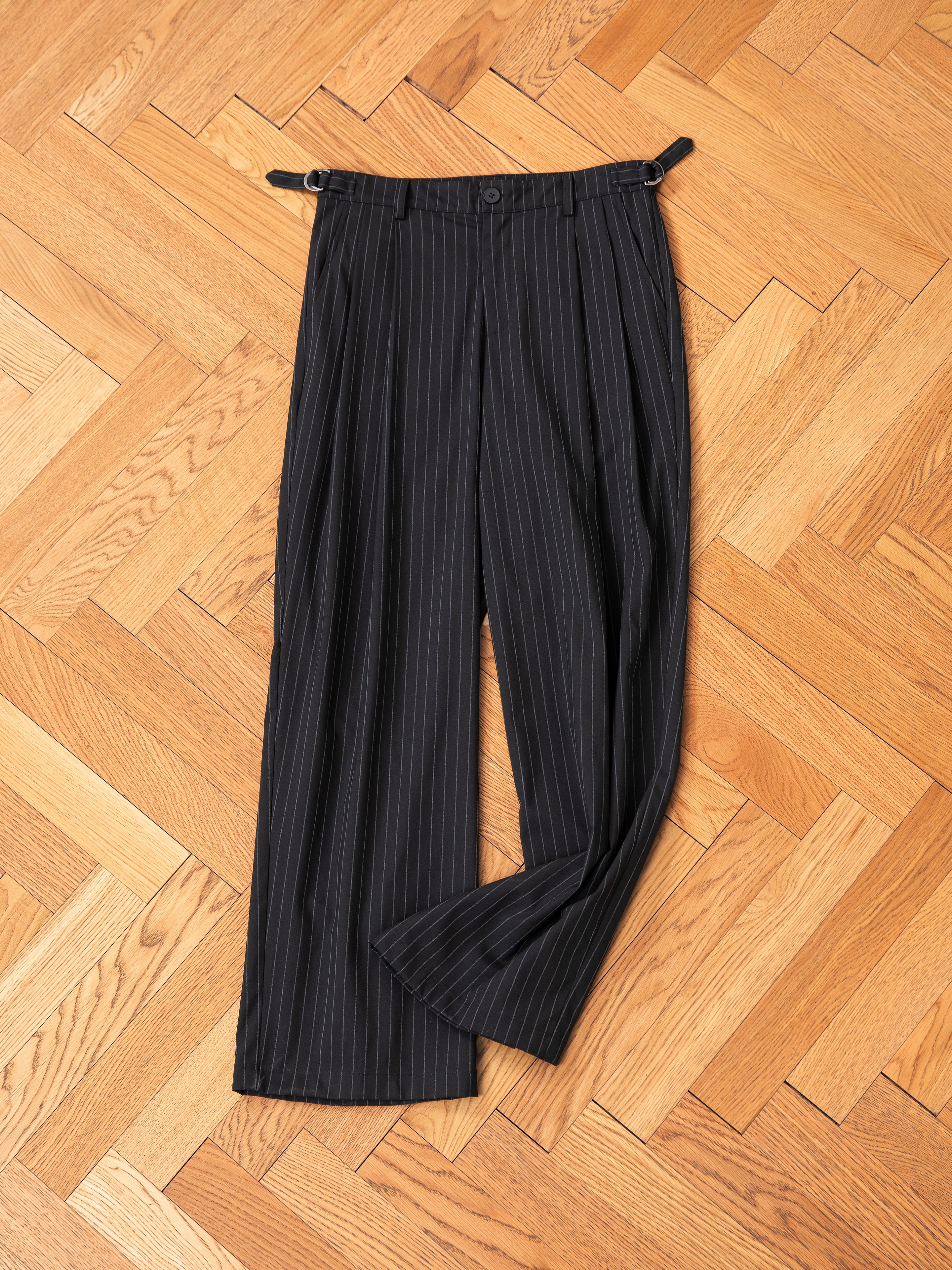 FOUND's Pinstripe Pleated Trousers, featuring a high-rise fit with wide legs and adjustable side buckles, rest elegantly on a wooden parquet floor.