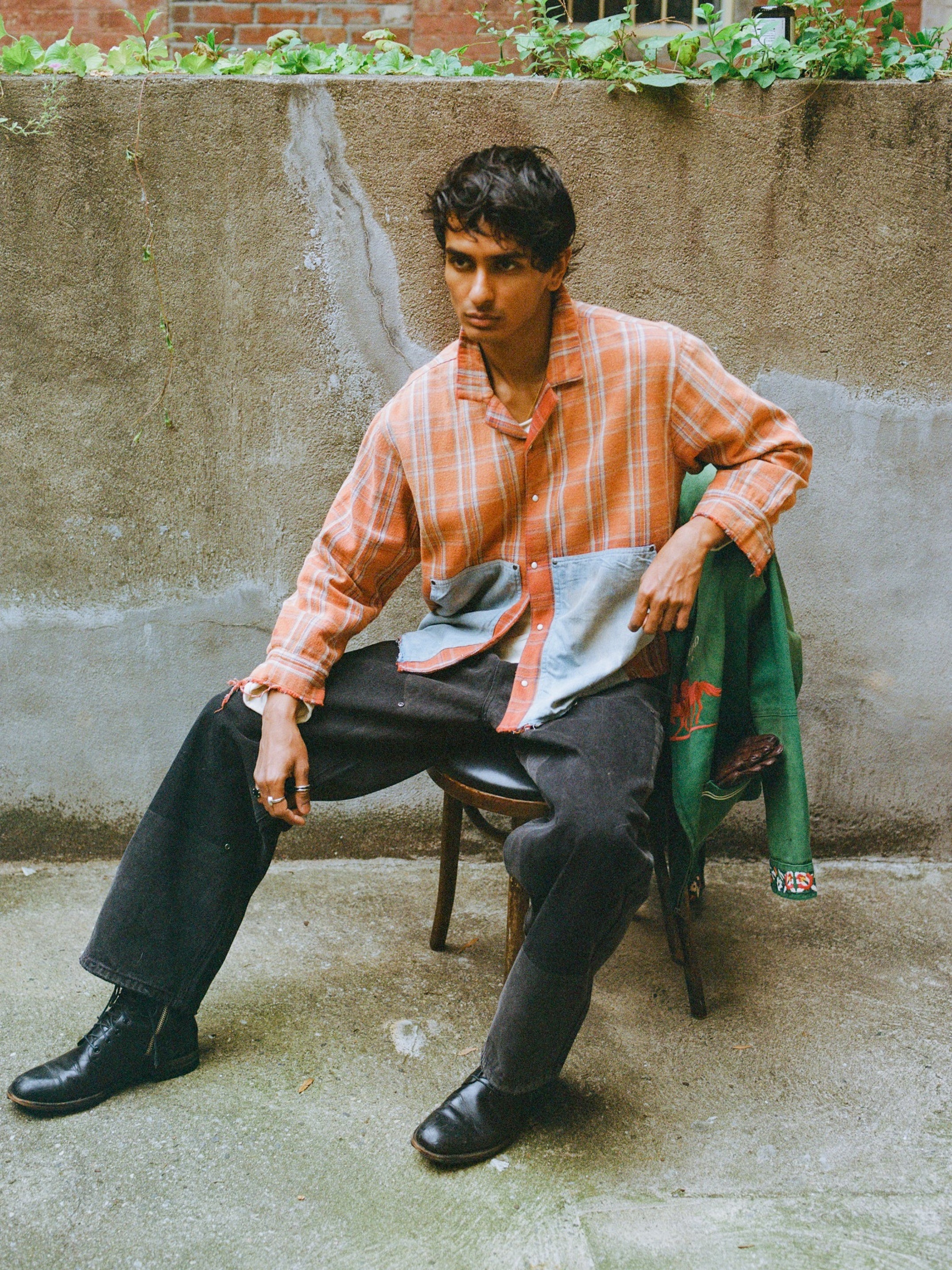 A man wears the FOUND Plaid Denim Shirt and black pants, sitting on a wooden chair outdoors against a concrete wall with a green jacket beside him, showcasing classic unisex workwear style.