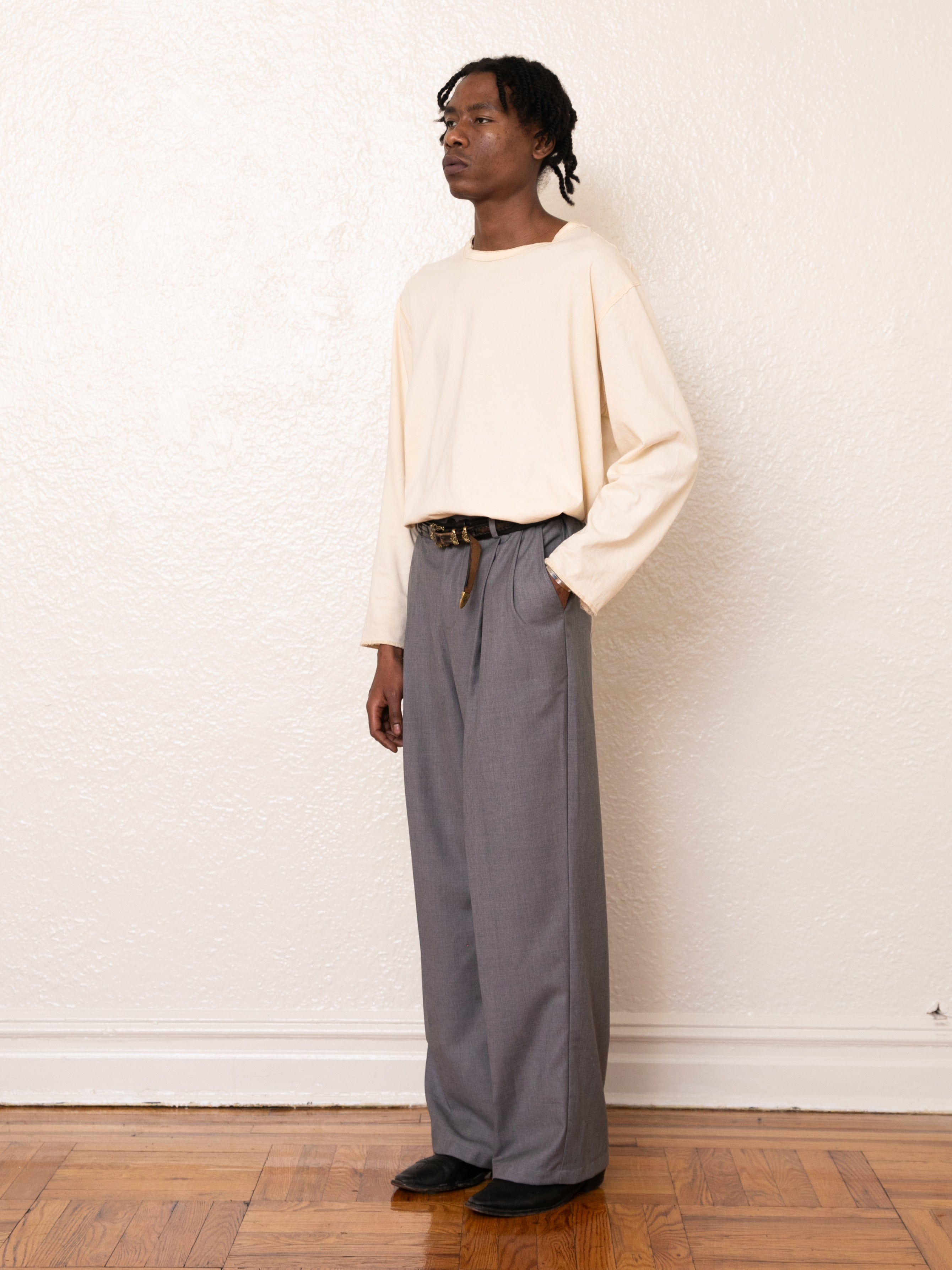 Someone stands against a cream wall, dressed in a white long-sleeve shirt, gray FOUND Pleated Trousers, and black shoes, with hands in pockets on a wooden floor.