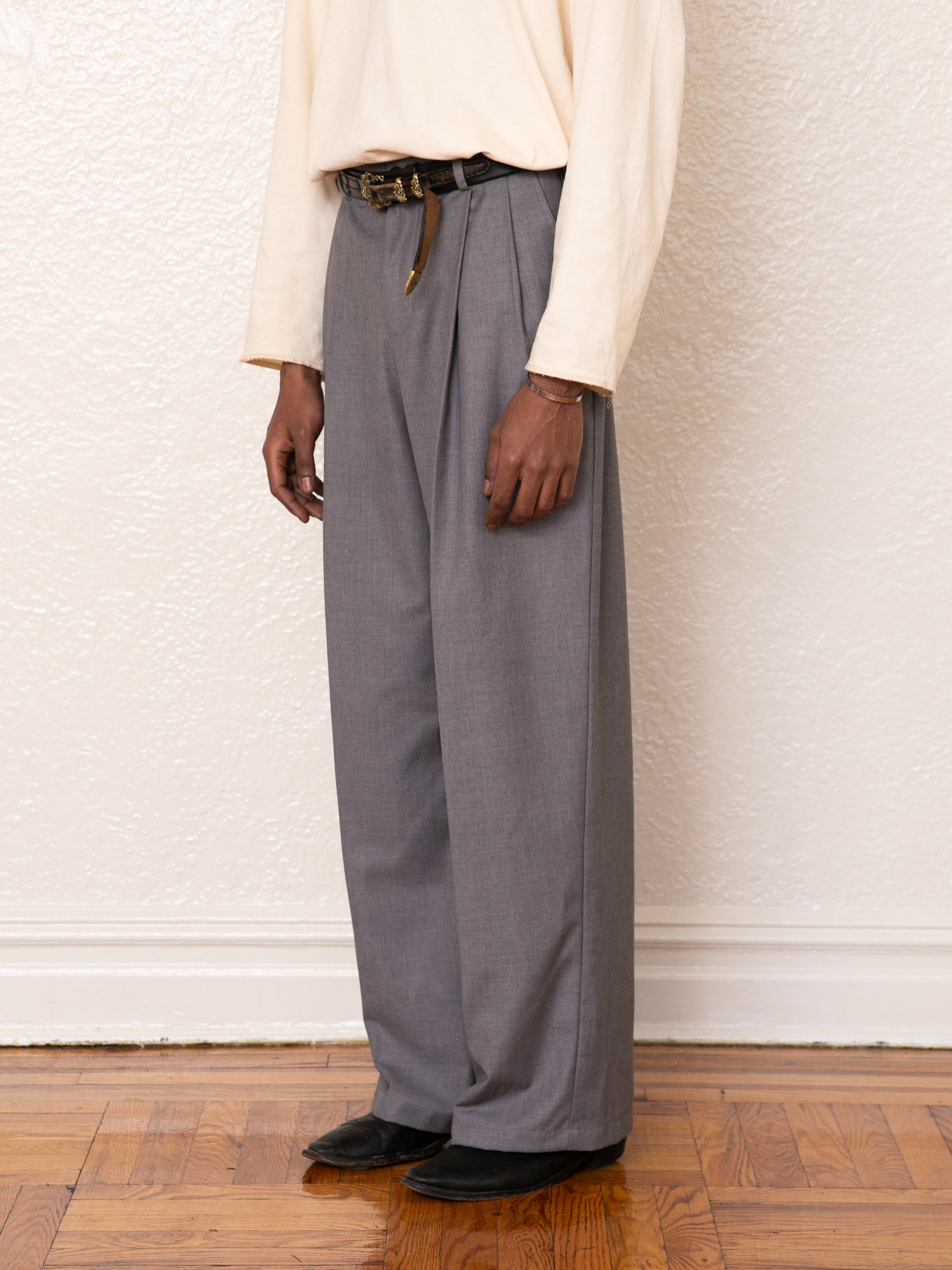 A person stands against a textured white wall and wooden floor, wearing FOUND Pleated Trousers in gray high-rise style with double pleats, paired with a beige long-sleeve shirt, black belt, and black shoes.