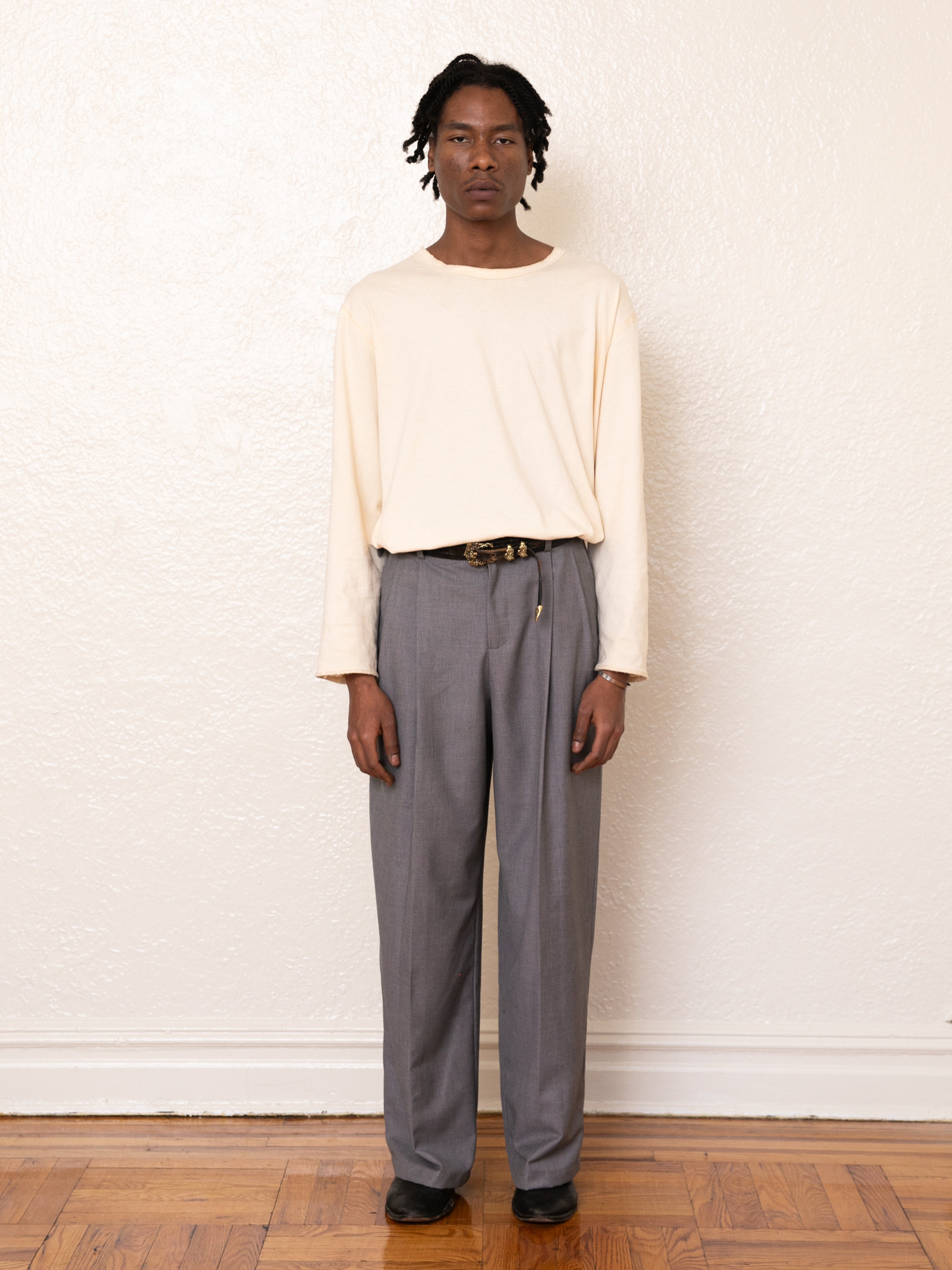 A person in a cream long-sleeve shirt, high-rise double pleated trousers by FOUND, and black shoes stands against a plain white wall on a wooden floor.