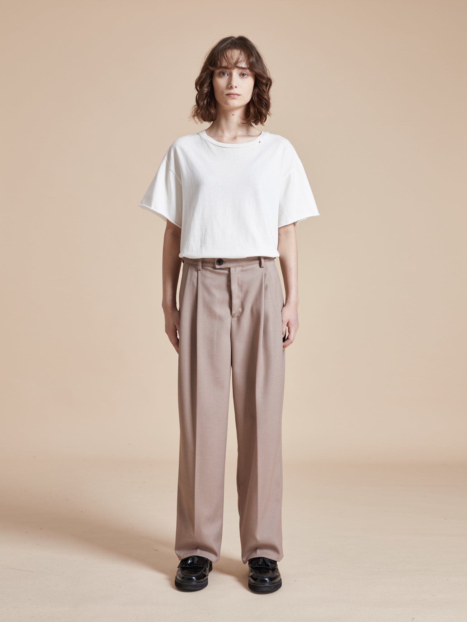 The model is wearing a white t-shirt and beige Found Pleated Trousers.