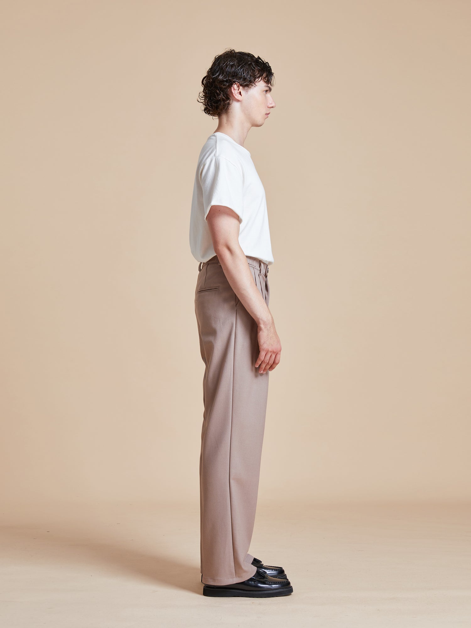 A man in a tan shirt and Pleated Trousers by Found is standing in front of a beige wall.