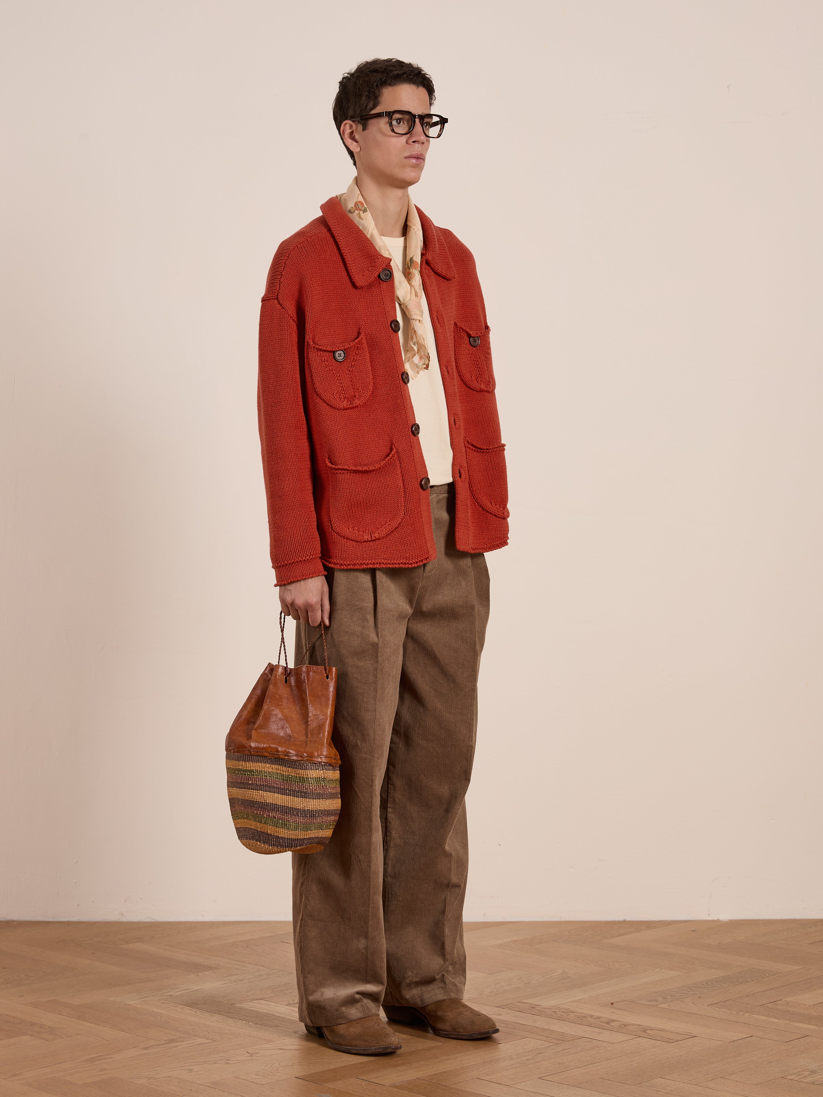 A person stands wearing a Hunter Knit Collared Cardigan by FOUND with sunglasses, styled with a striped shirt. They pair the outfit with brown pants and a striped bag, all against a wooden floor and plain background—perfect for transitional layering.