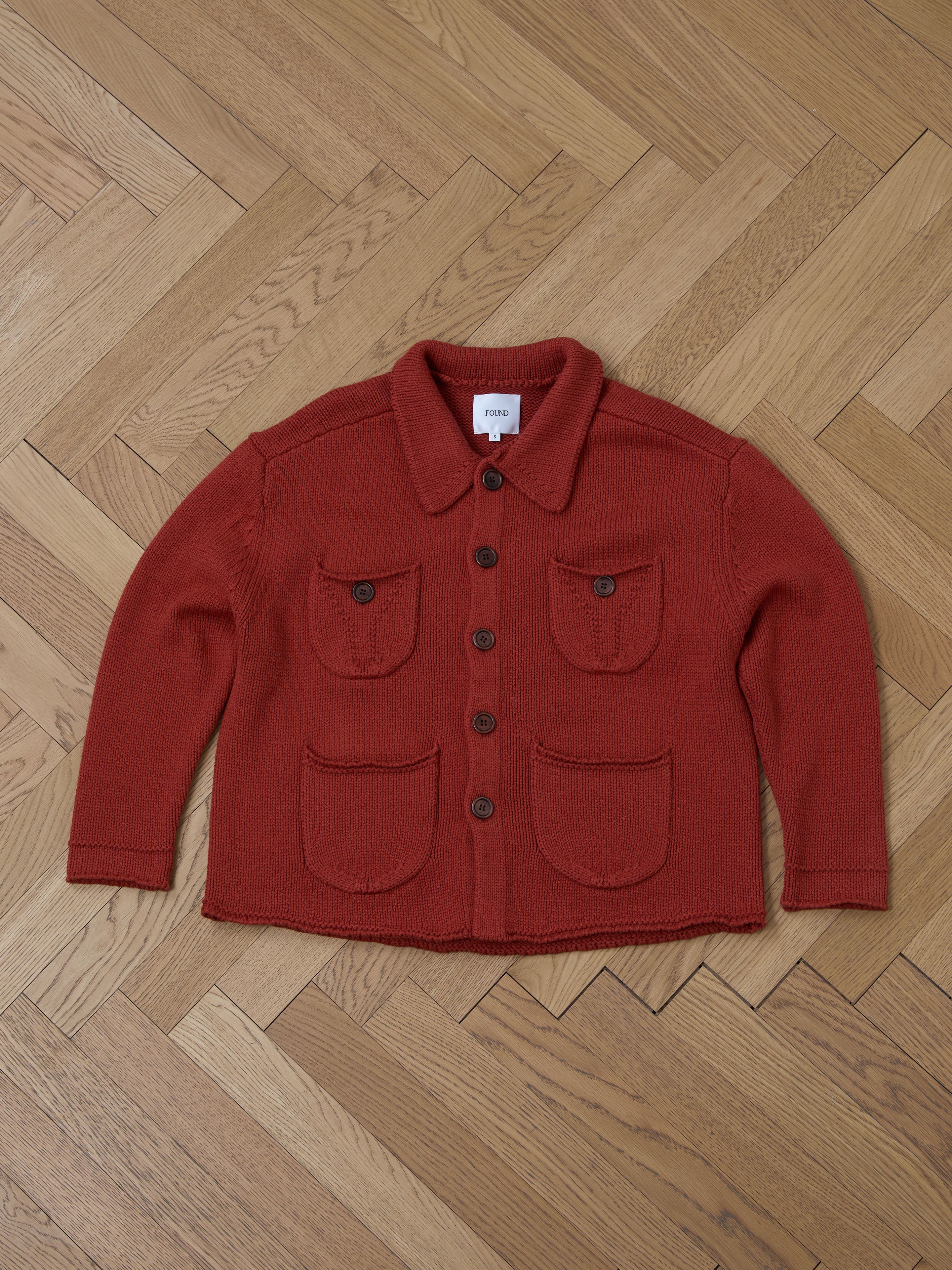 The Hunter Knit Collared Cardigan by FOUND, designed for transitional layering, is a red heavy knit cotton piece with a collar and four front pockets, showcased on a wooden herringbone-pattern floor.