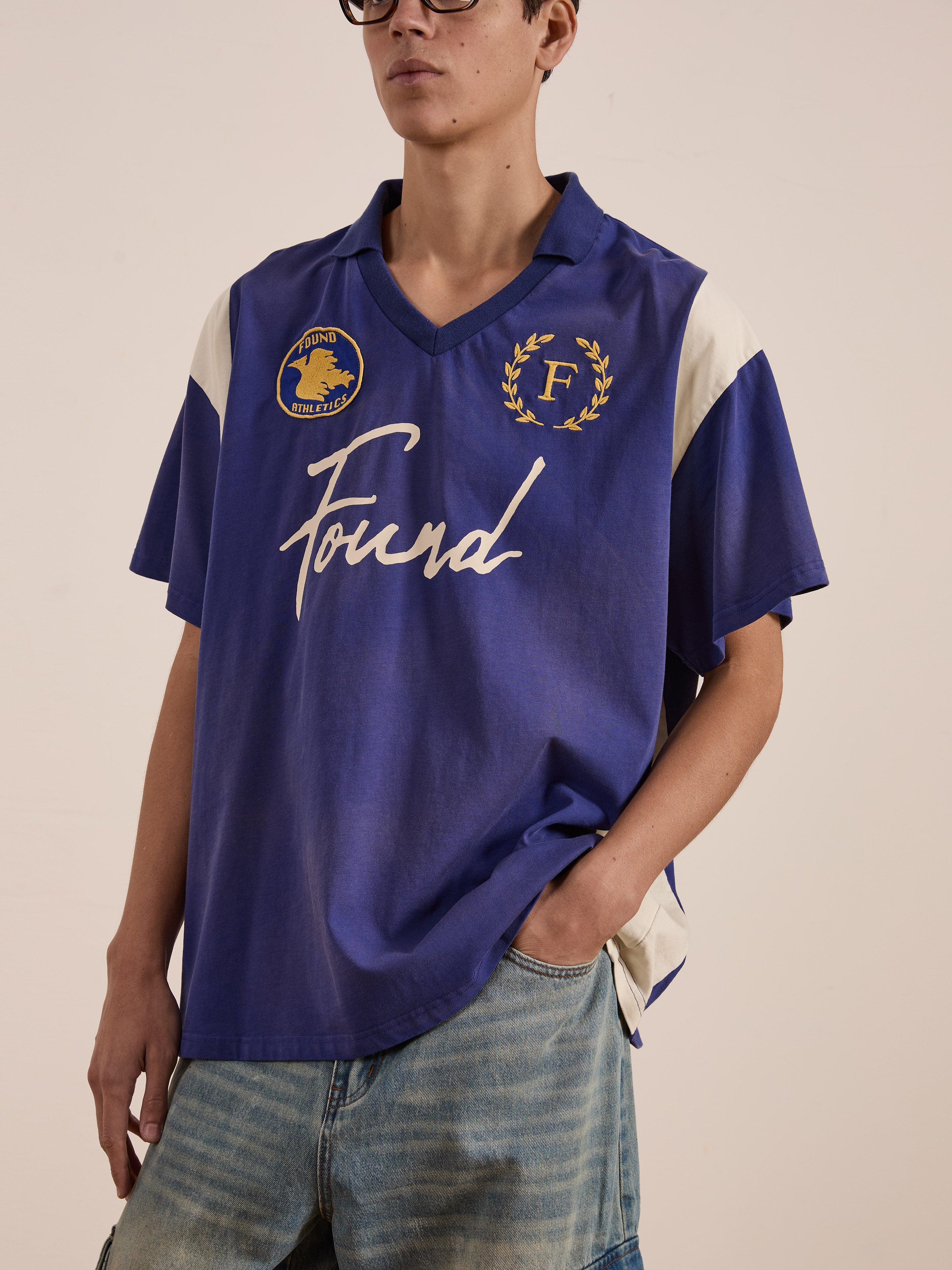 Sporting a vintage sportswear vibe, someone is wearing the FOUND brand's Premier League Jersey in blue, adorned with an embroidered crest and various emblems. Paired with light blue jeans, this look exudes casual chic style.