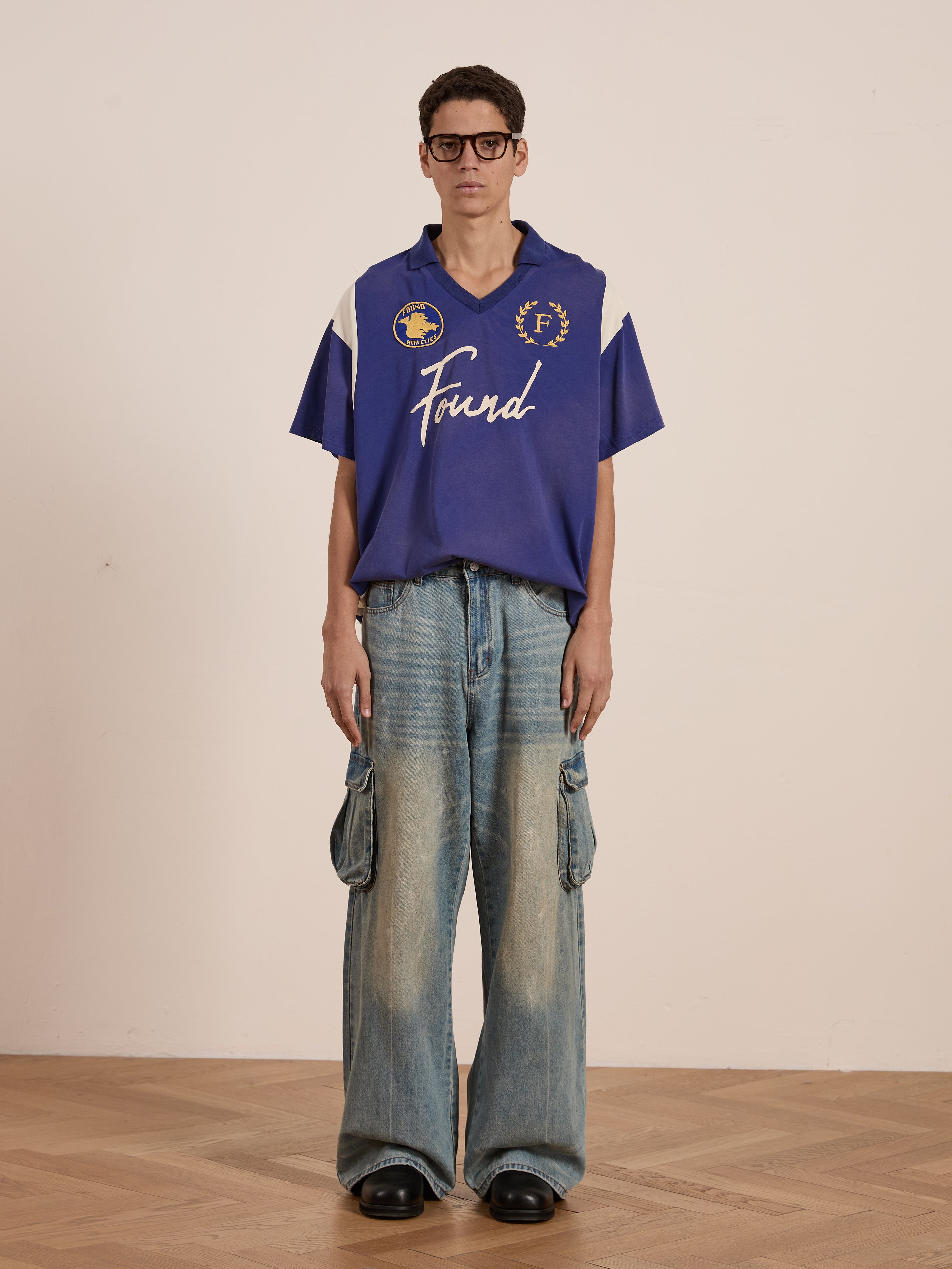 A person wearing glasses stands on a wooden floor against a plain backdrop, dressed in wide-legged jeans and a blue Premier League Jersey by FOUND, featuring an embroidered crest.