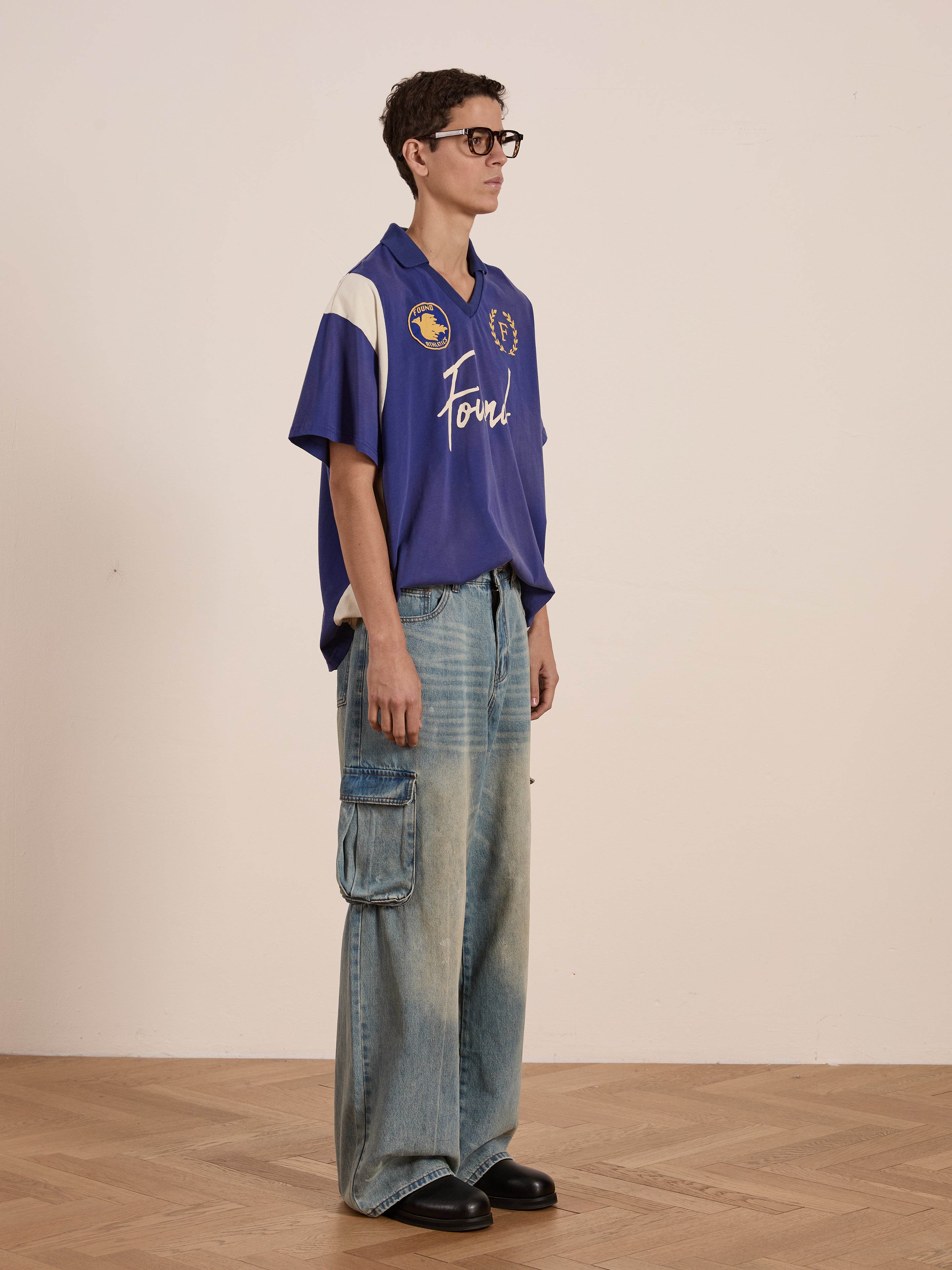 A person in a blue Premier League Jersey by FOUND and wide denim cargo pants stands on a wooden floor against a plain background, subtly channeling vintage sportswear vibes reminiscent of an embroidered crest design.