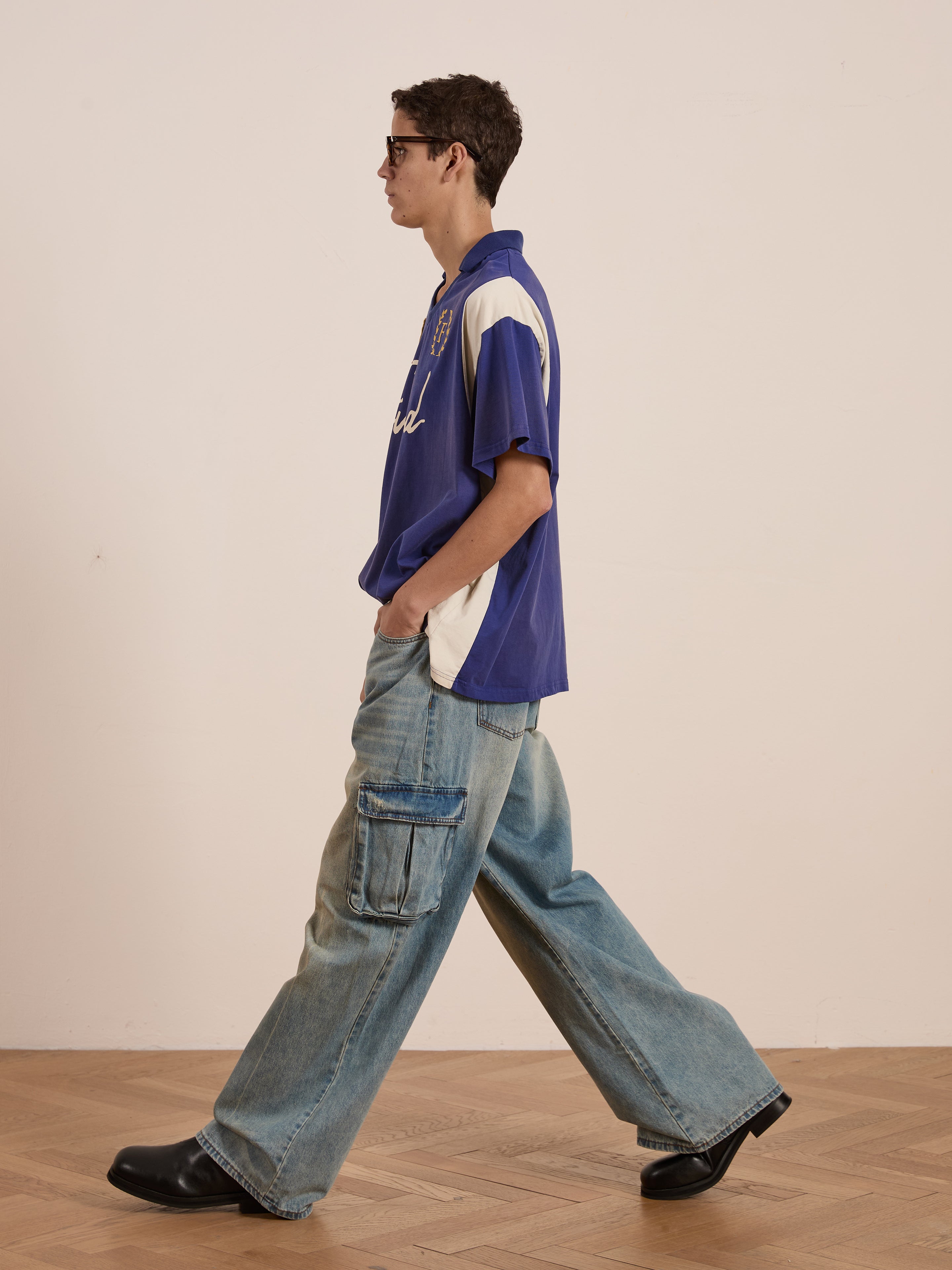 A person in sunglasses walks sideways, exuding a vintage sportswear vibe with a blue and white FOUND Premier League Jersey, wide-legged cargo jeans, and black shoes on a wood floor.