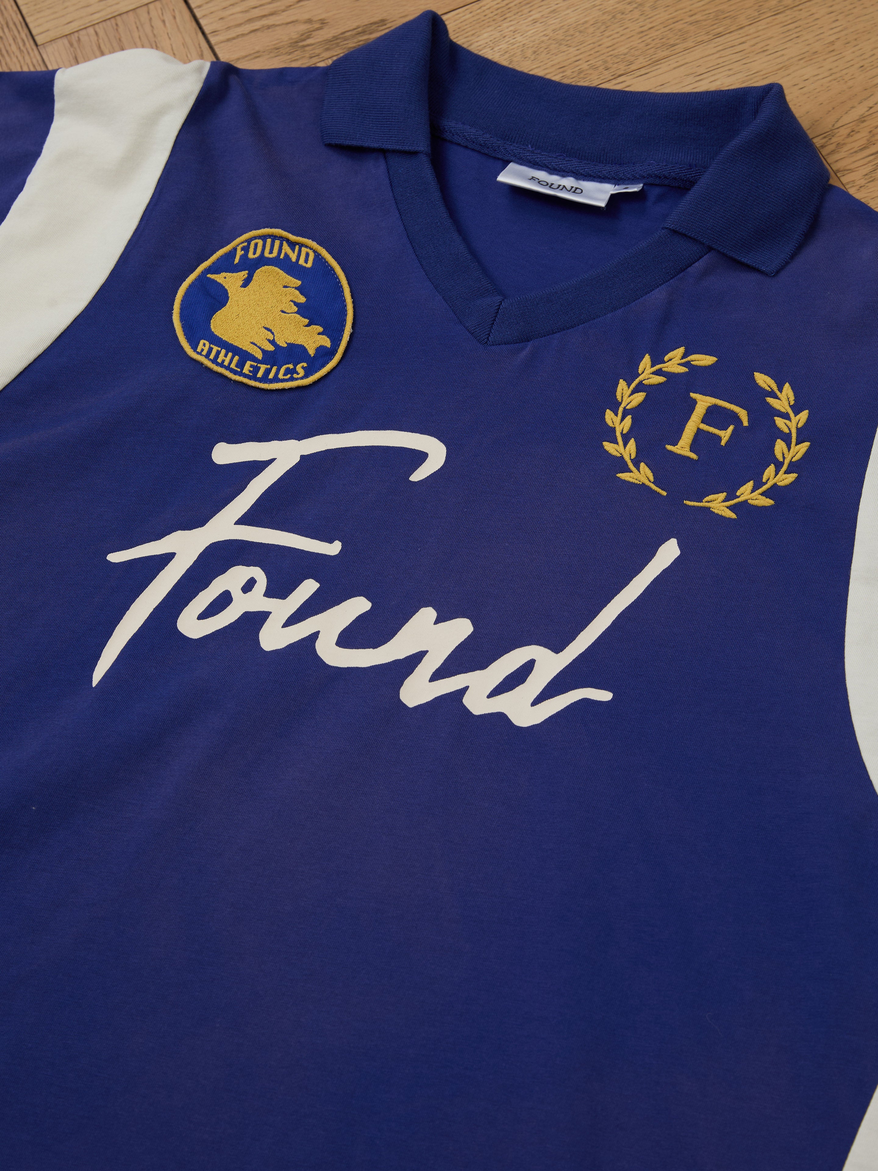 The Premier League Jersey by FOUND is a blue and white vintage sportswear piece featuring the "Found" logo, an "F" with a laurel, and an embroidered crest of a running figure with "Found Athletics." Ideal for vintage sportswear enthusiasts.