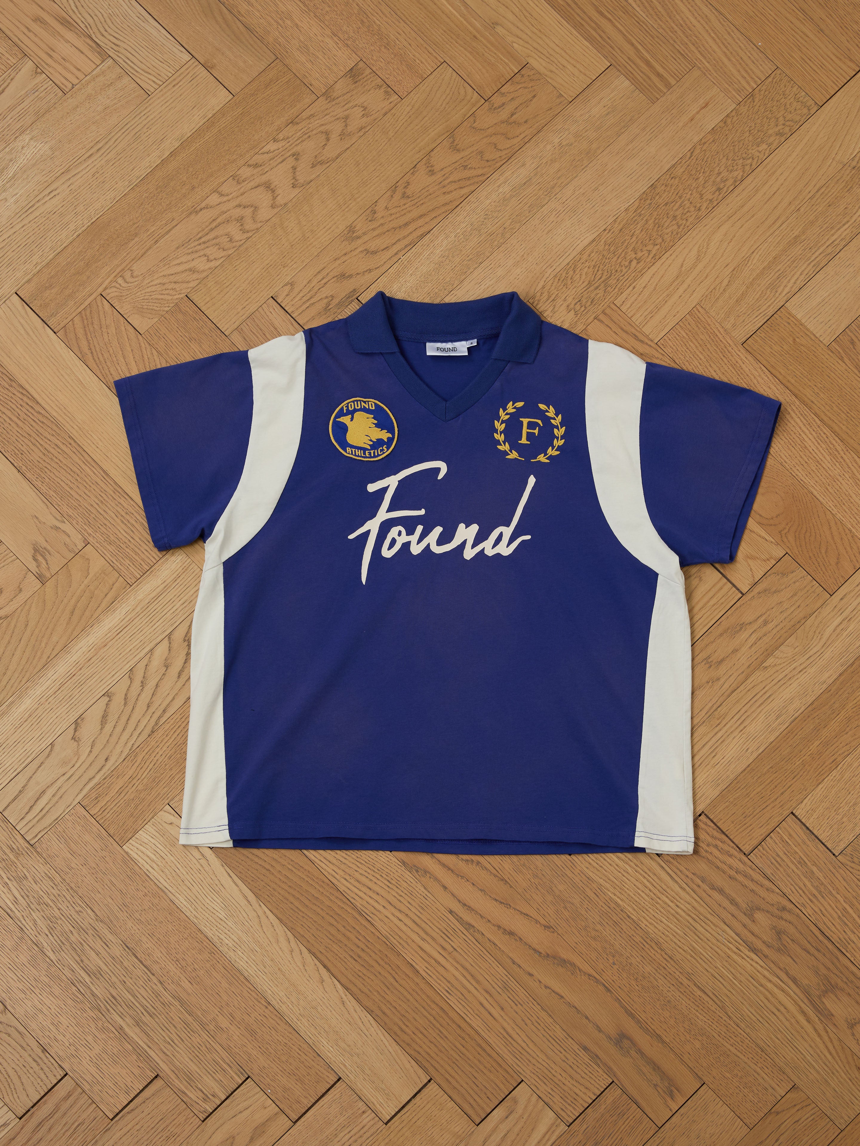 A blue and white Premier League Jersey from FOUND, with short sleeves, logos, an embroidered crest, and "Found" on the front, displayed on a wooden floor.