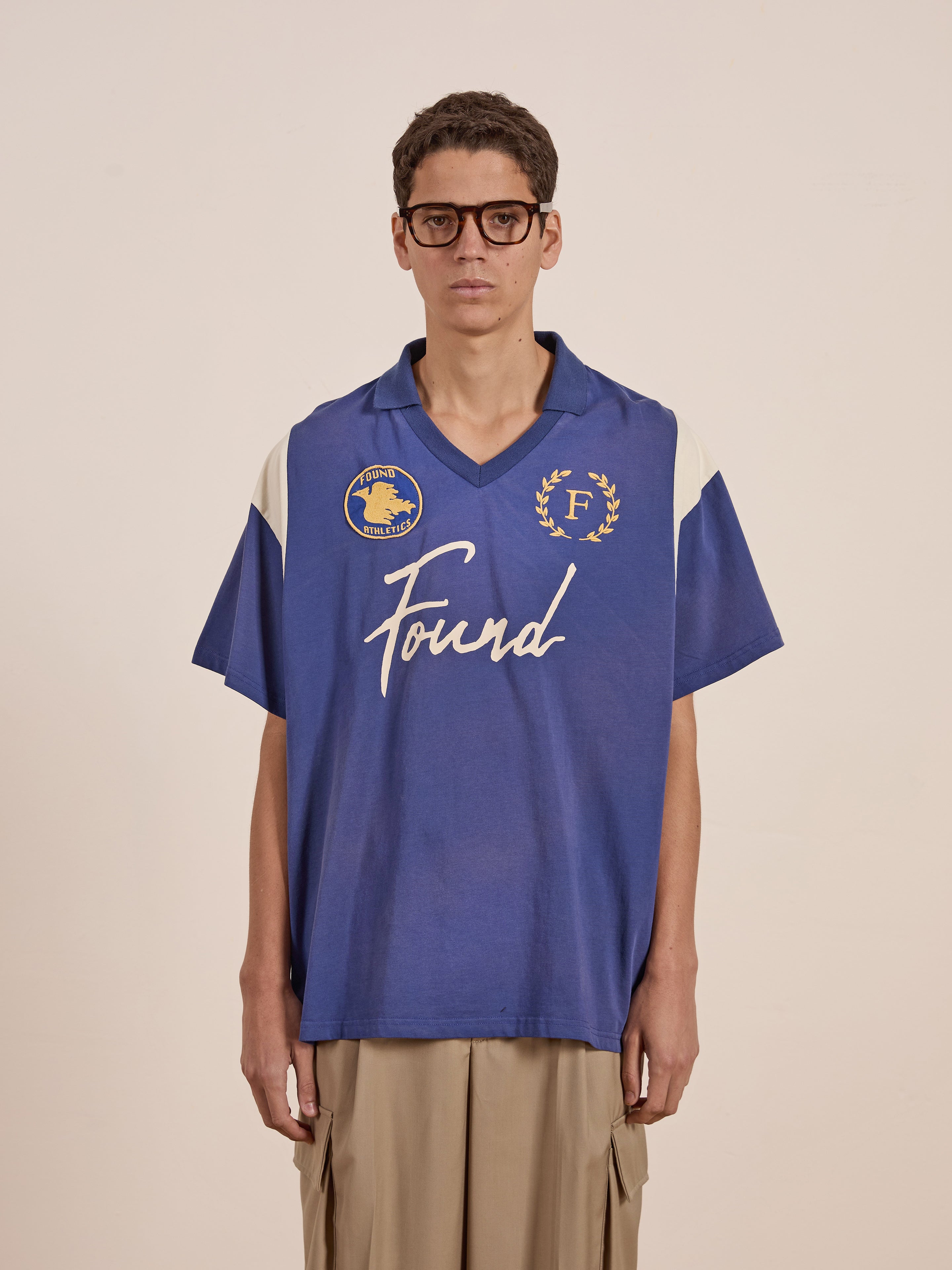 A person in glasses wears a blue oversized shirt featuring vintage sportswear logos and "FOUND" branding, paired with beige pants, against a plain background.