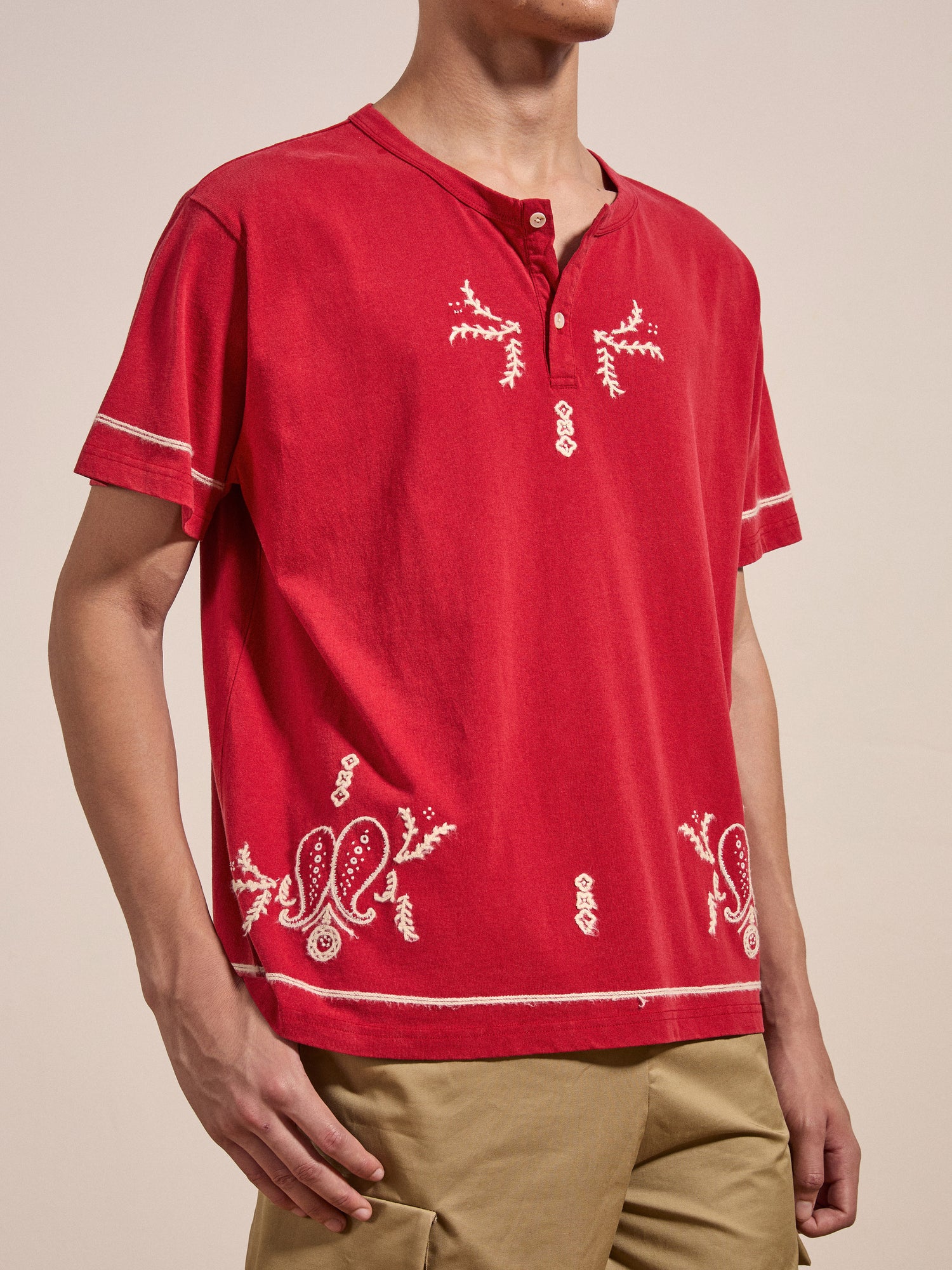 A person is wearing an Embroidered Henley by FOUND, which is a red short-sleeve shirt with white embroidery and a round neck with buttons, paired with beige pants.