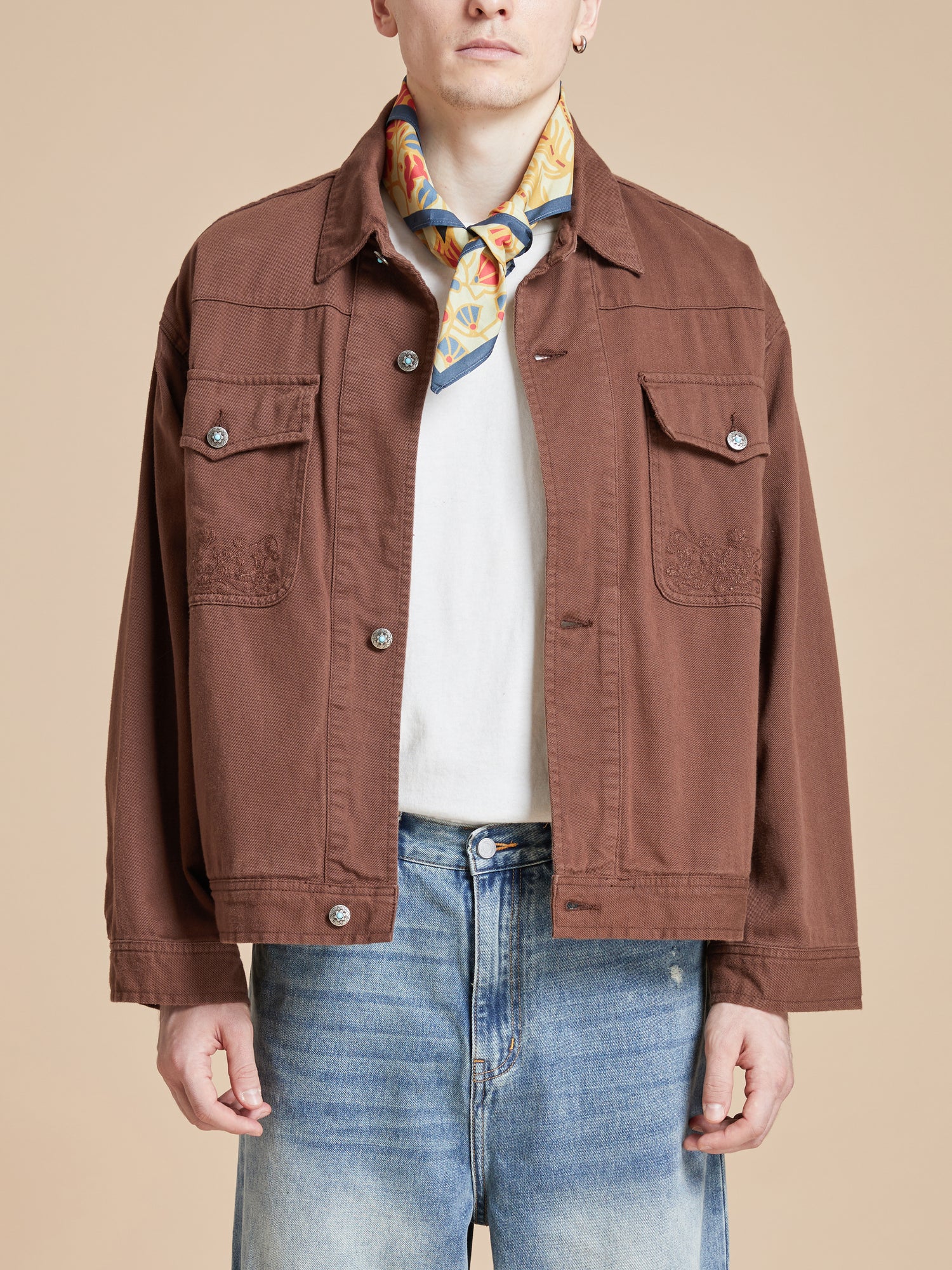 A man wearing a brown jacket and jeans, accessorized with the Coming Soon Rainforest Bandana by Found.