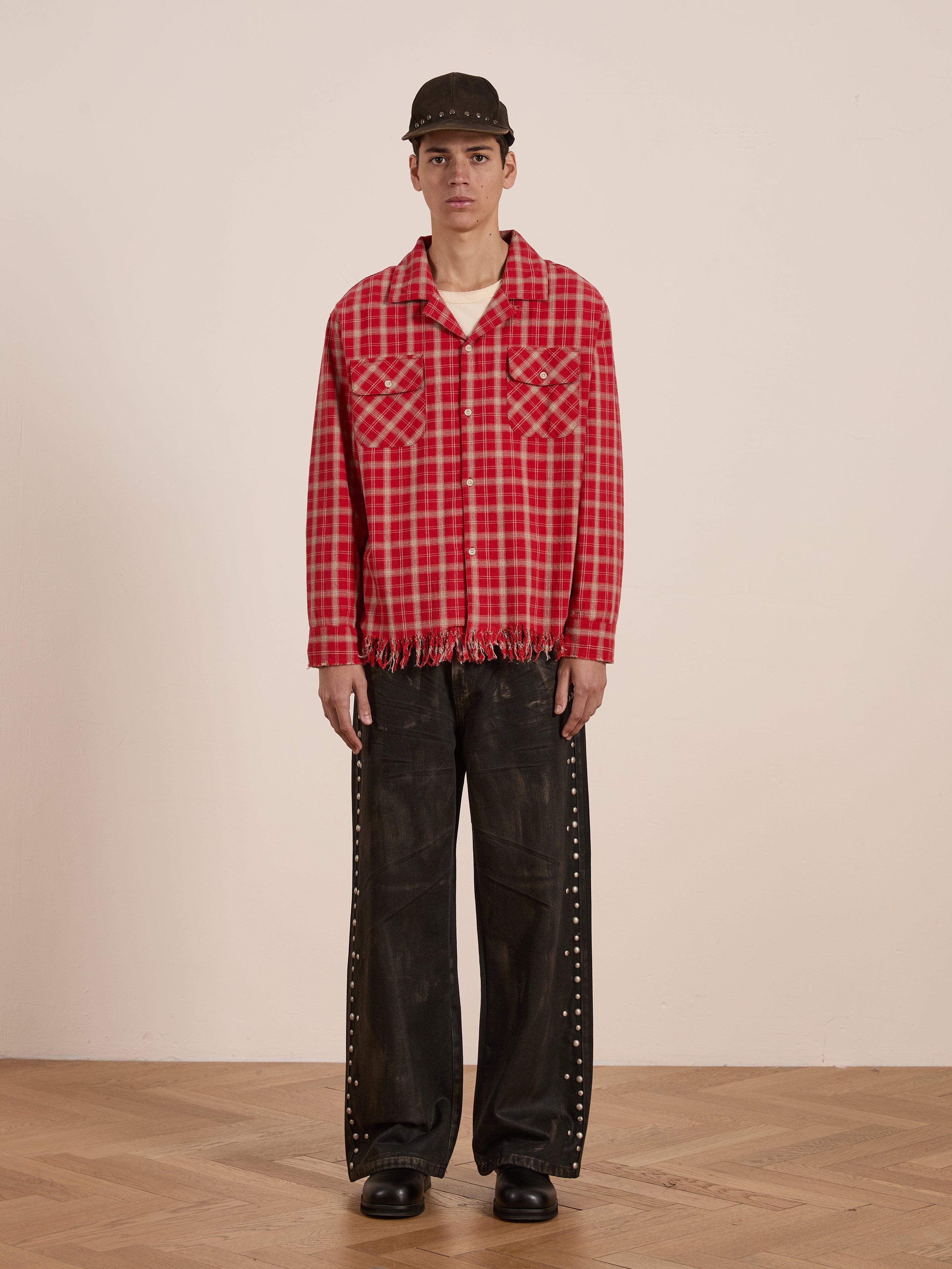 Individual dressed in a Raw Cut Fringe Plaid Camp Shirt by FOUND, paired with a black cap, black wide-leg pants featuring stud accents, and black shoes, posed against a simple backdrop.