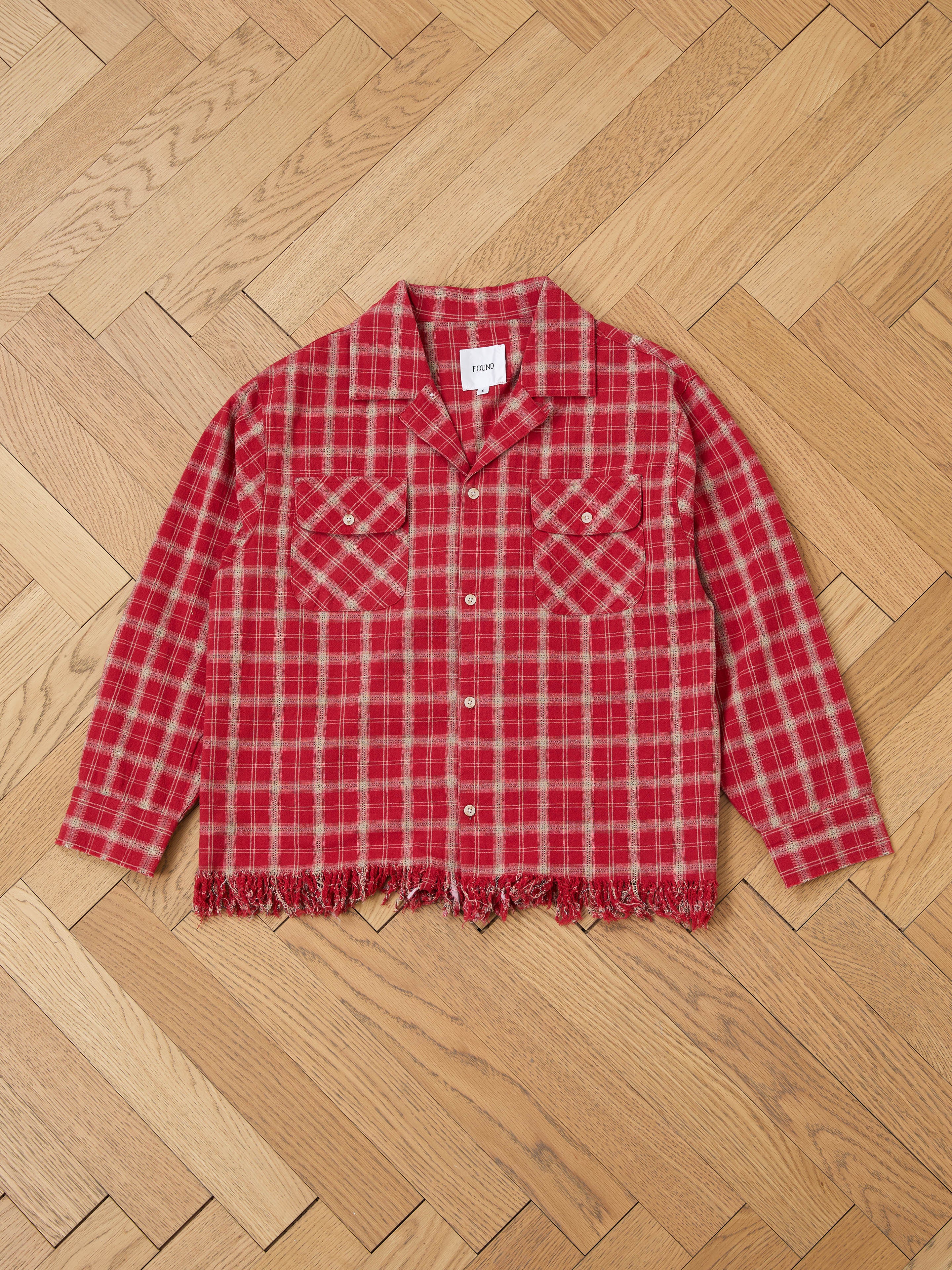 The Raw Cut Fringe Plaid Camp Shirt by FOUND is a cozy red flannel with two chest pockets and a frayed hem, showcased on a wooden herringbone floor.