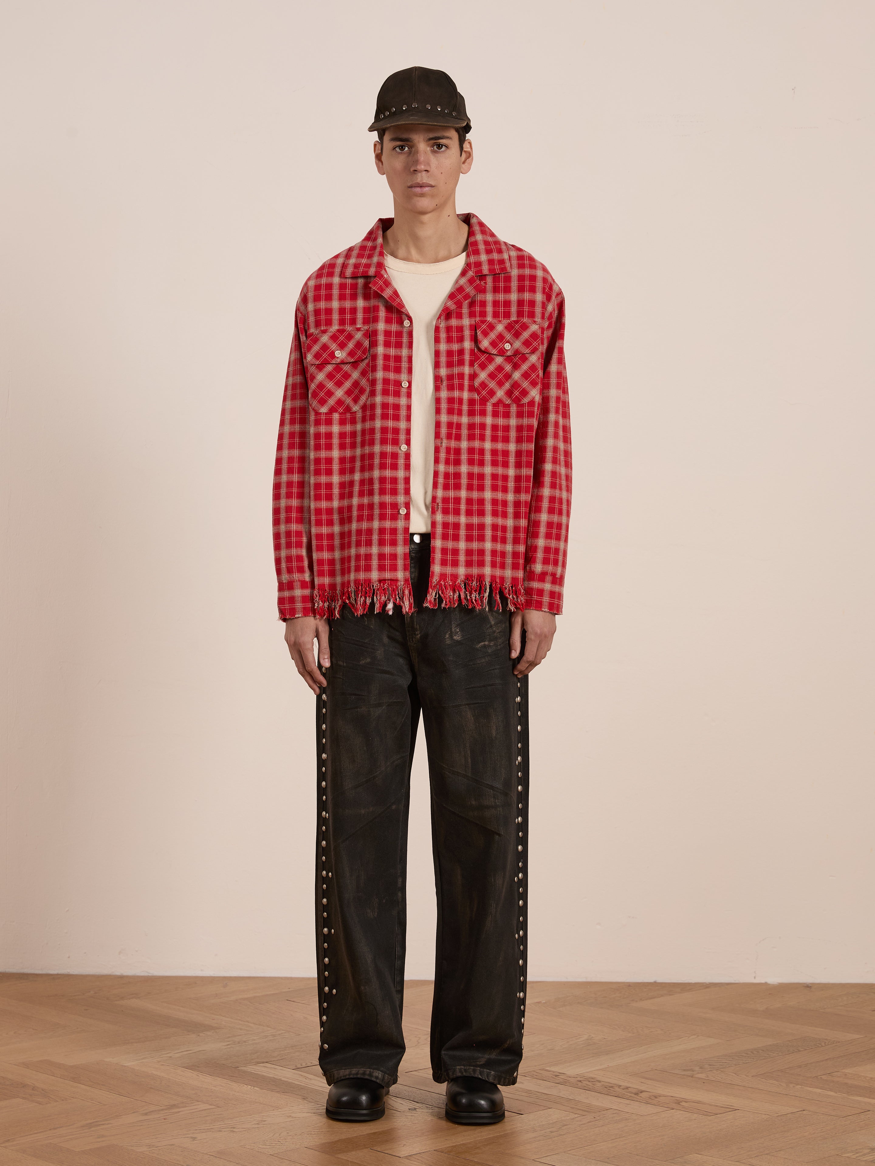 A person indoors wears a red Raw Cut Fringe Plaid Camp Shirt from FOUND, paired with studded black pants, black shoes, and a black cap.