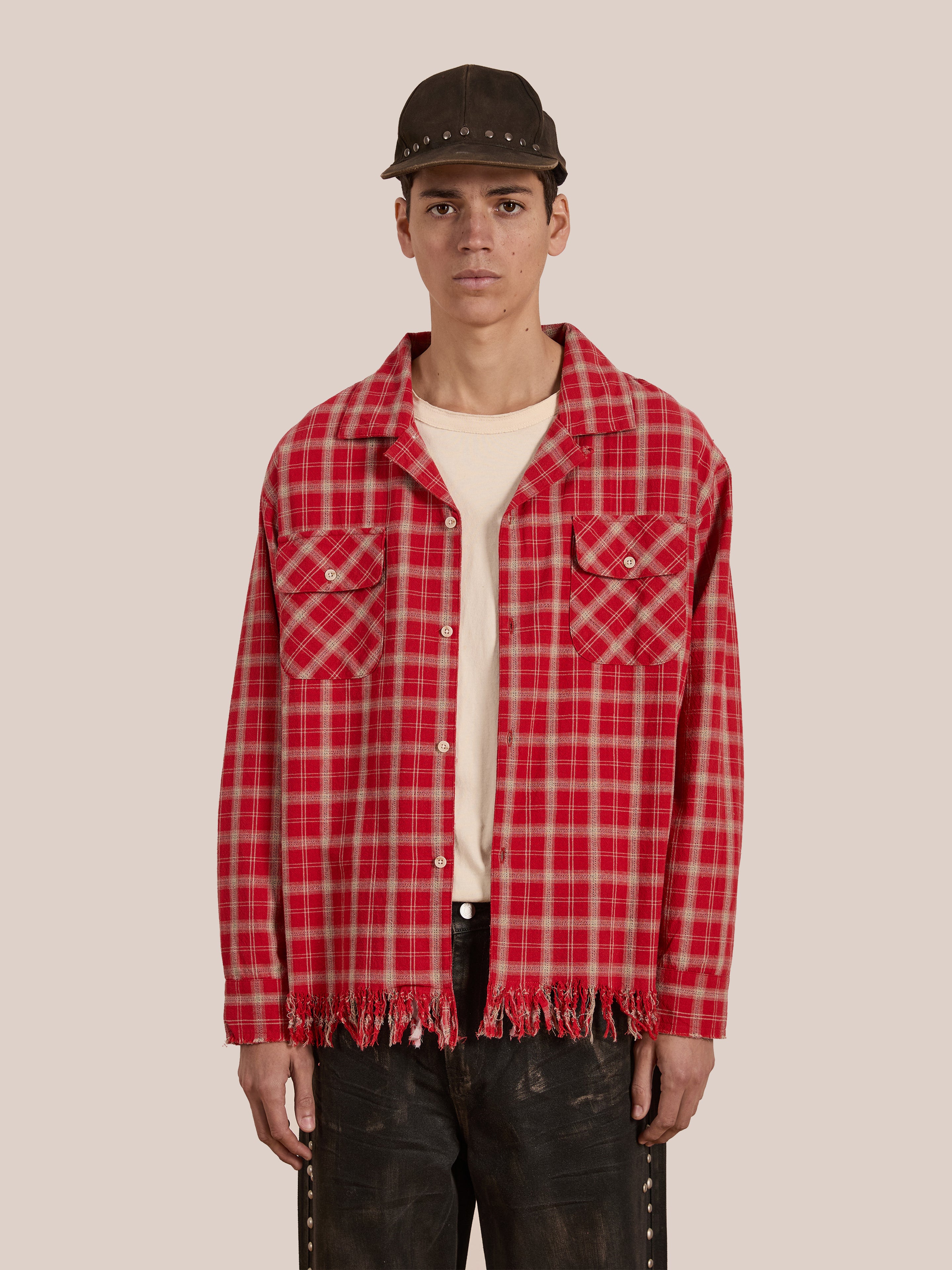 A person in a FOUND Raw Cut Fringe Plaid Camp Shirt with black pants, beige shirt underneath, and a black cap stands against a plain backdrop. The outfit exudes vintage Americana charm while ensuring flannel comfort.