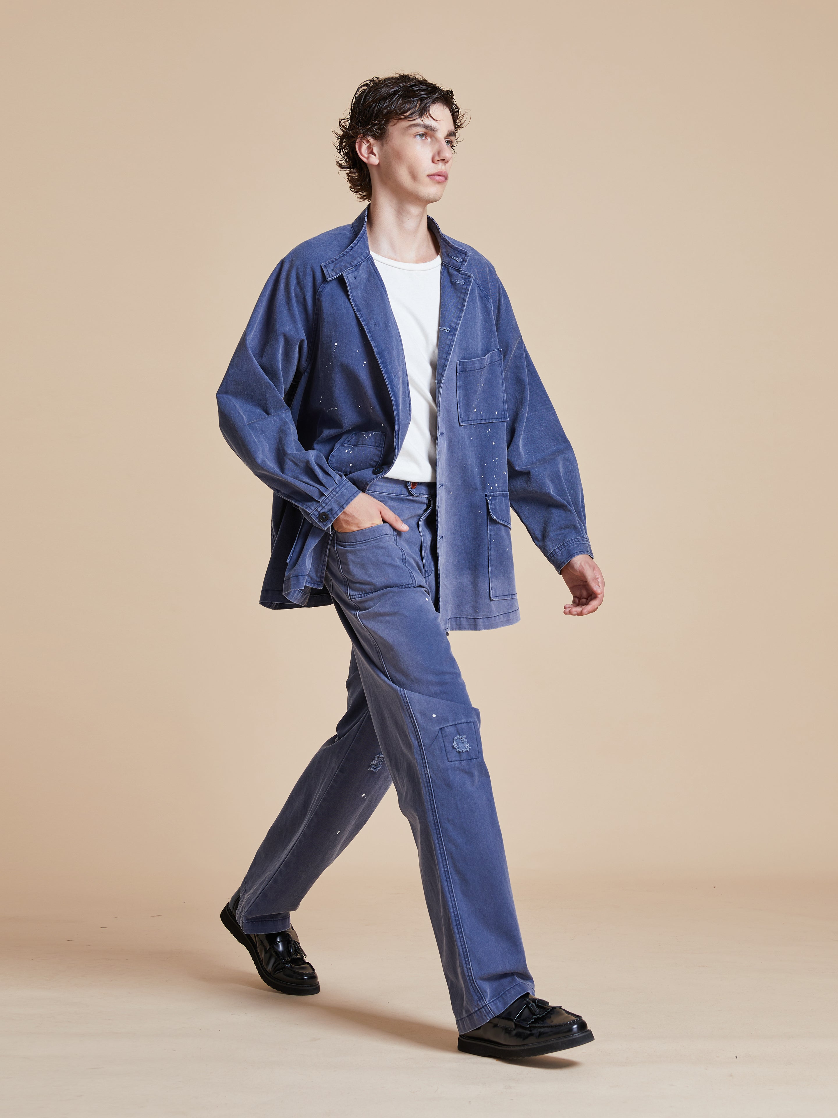 A man in blue pants walking on a beige background wearing the Found River Painters Chore Coat.