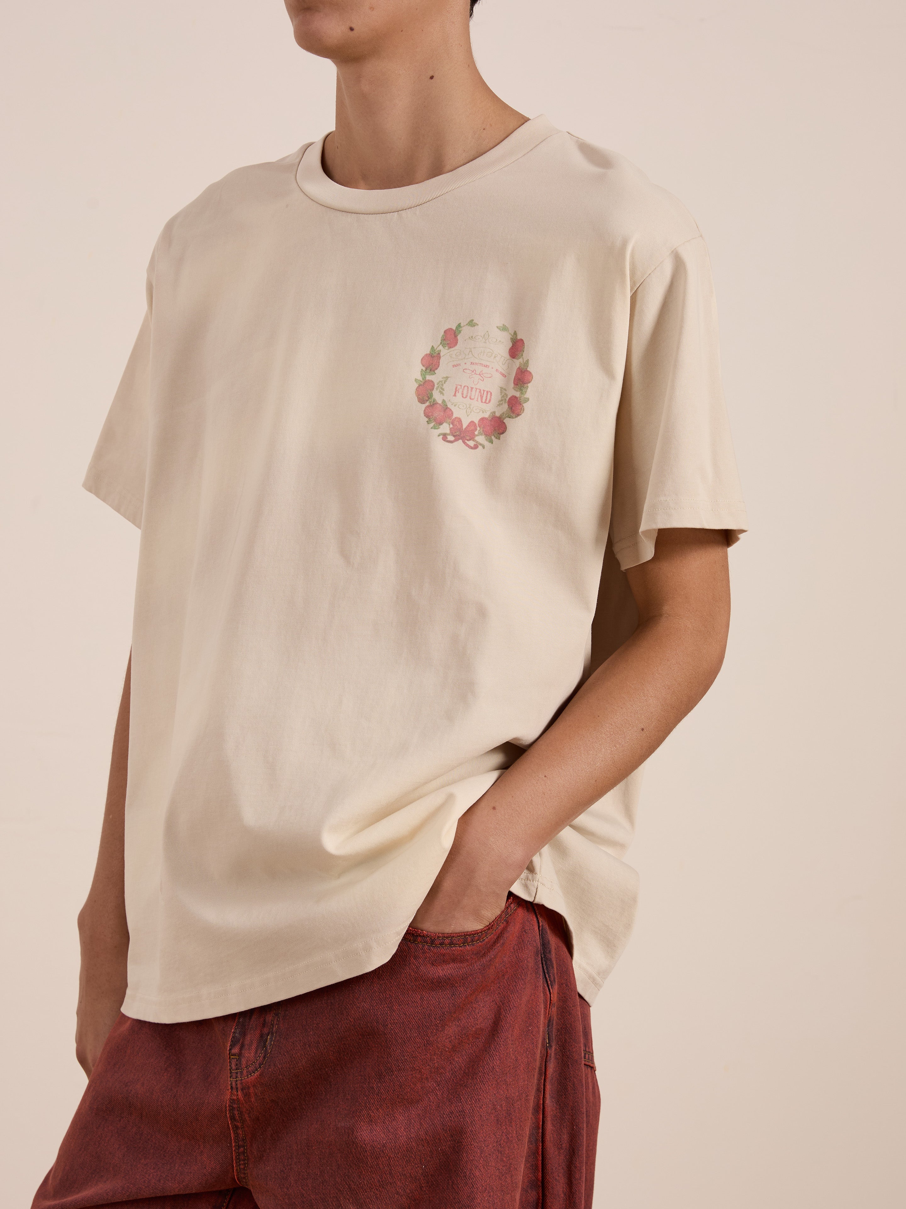 Individual in a cream FOUND Rose Garden Tee featuring a floral wreath graphic and faded text, matched with red pants.