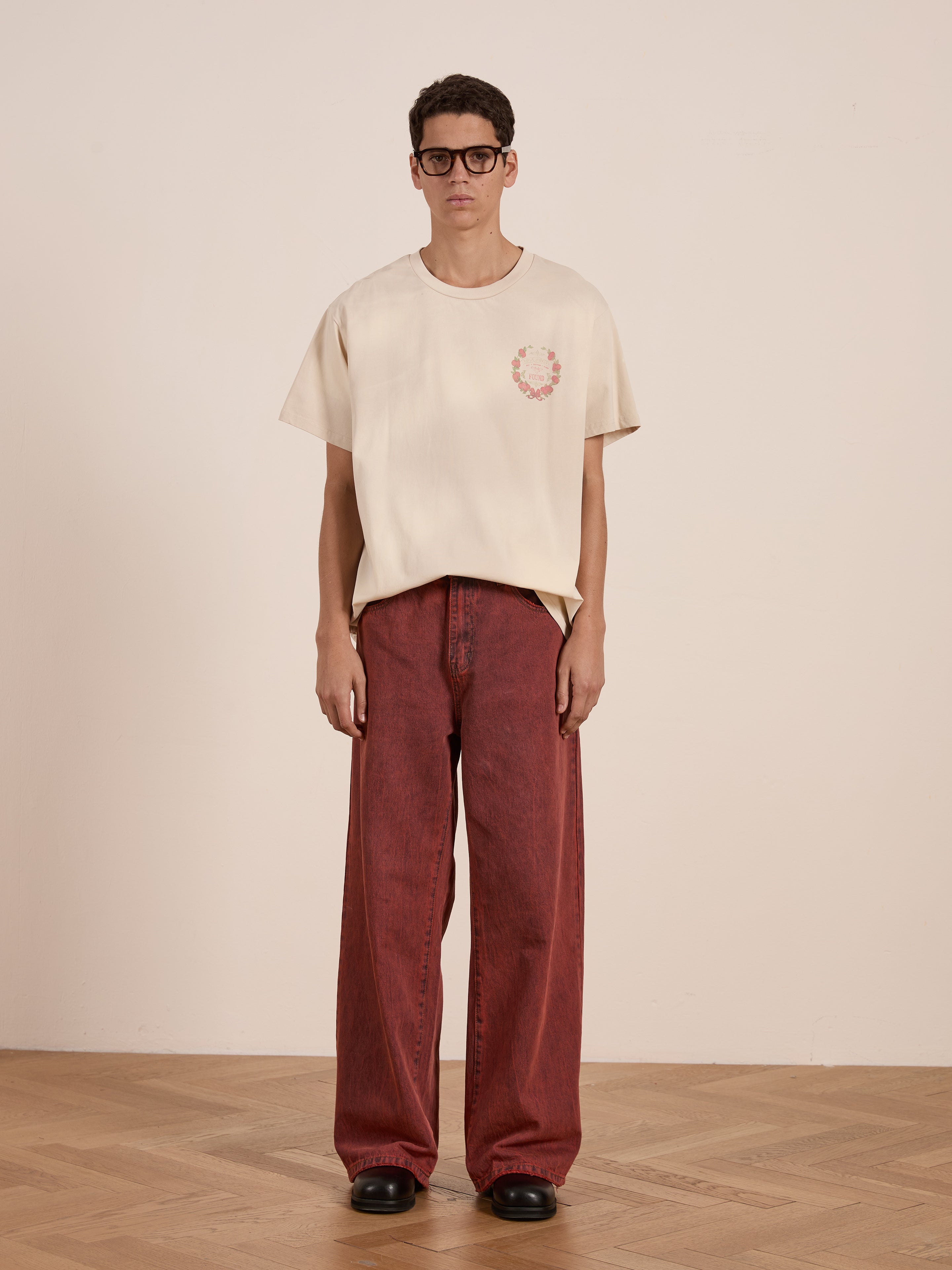 A person in FOUND's Rose Garden Tee in beige, featuring a small logo, paired with wide red jeans and black shoes, stands on a wooden floor against a plain wall, exuding vintage charm.