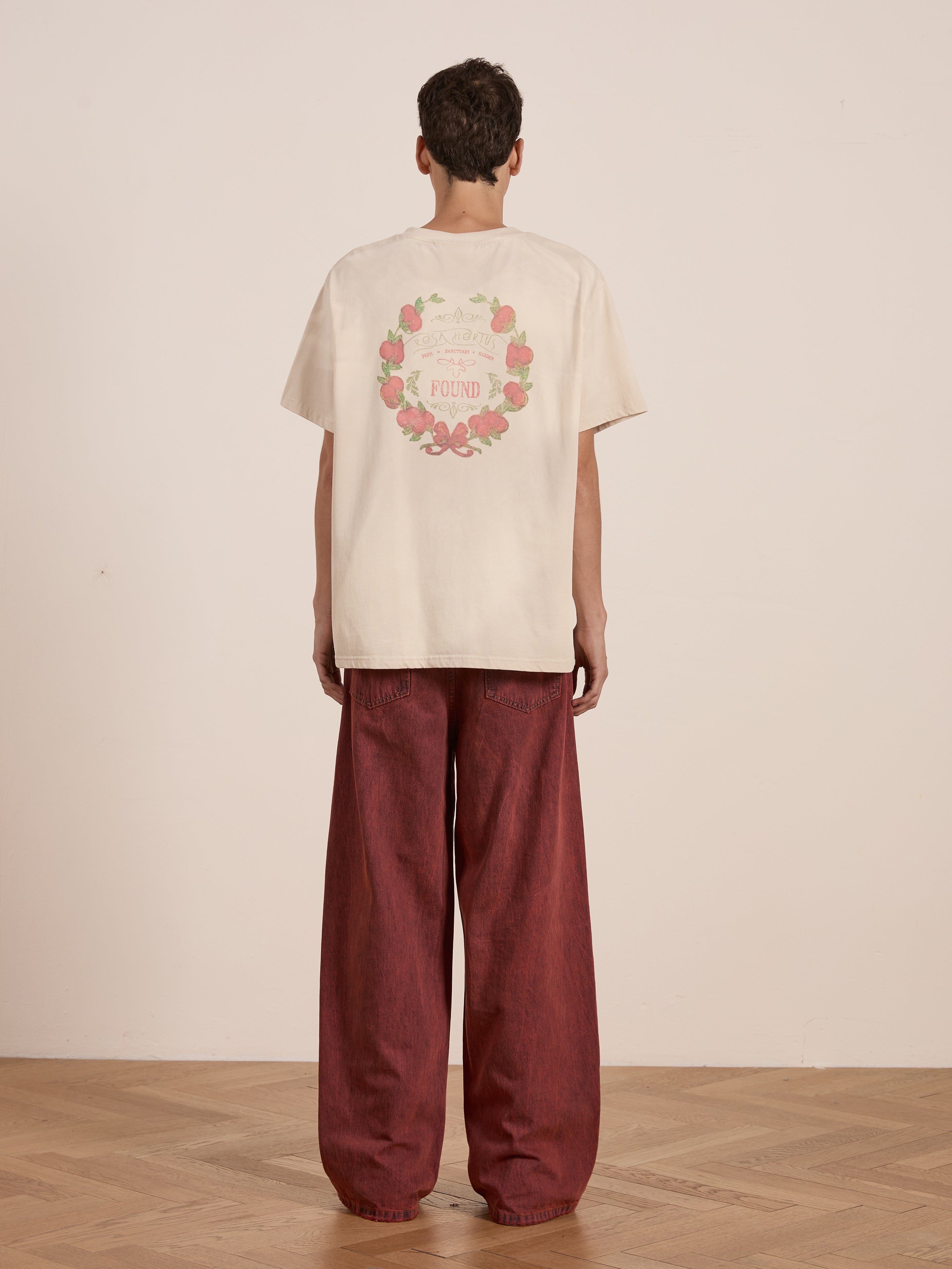 A person turns away, highlighting the vintage charm of FOUND's beige Rose Garden Tee with its floral wreath design, expertly paired with wide-legged red pants.