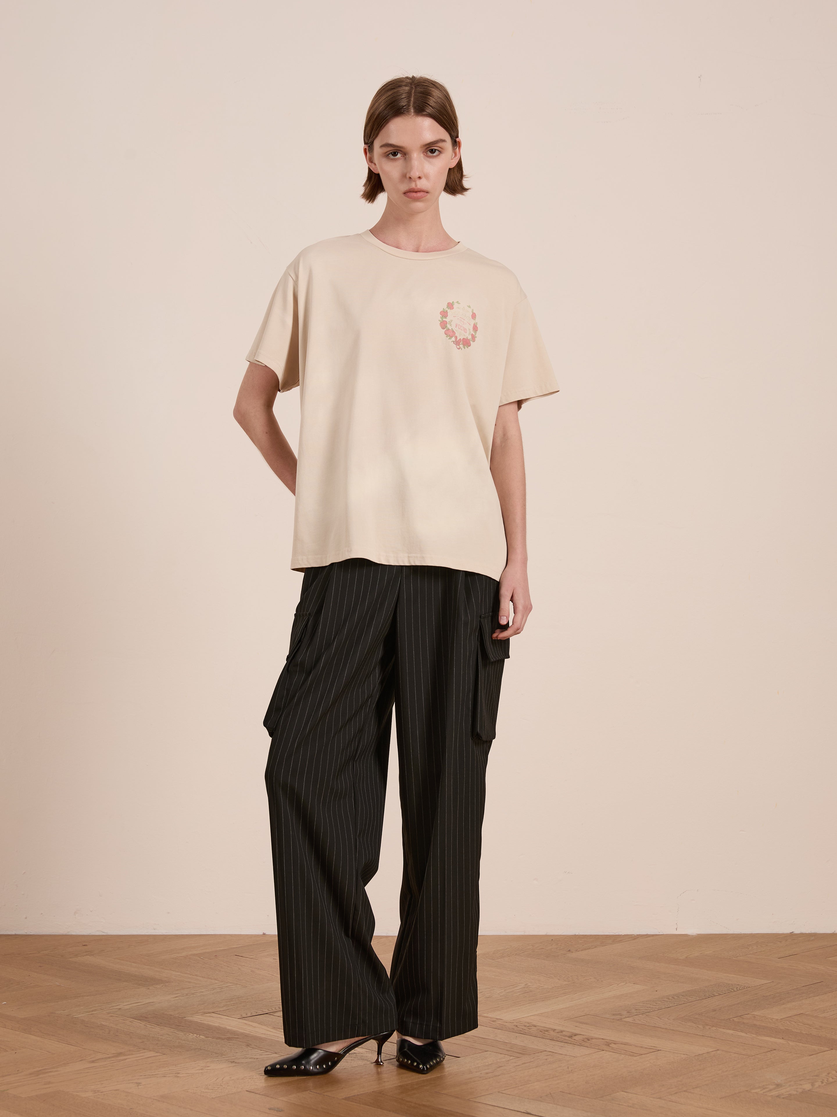 A person stands against a beige wall wearing the Rose Garden Tee by FOUND and dark pinstriped cargo pants.