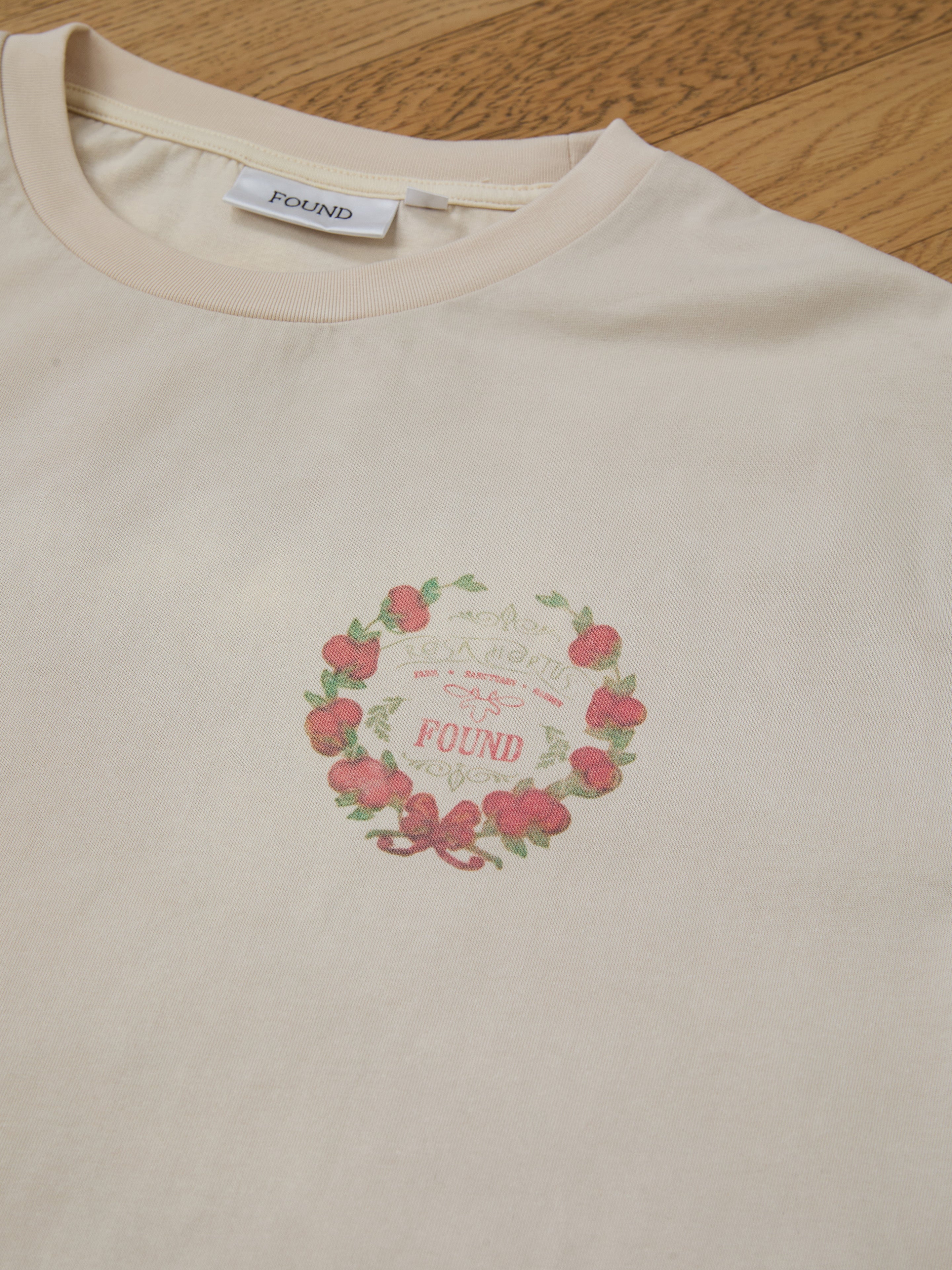 A beige T-shirt, the Rose Garden Tee by FOUND, features a vintage charm with a floral crest design. Red flowers elegantly wreath around "FOUND," embodying its essence as it rests on a wooden surface.