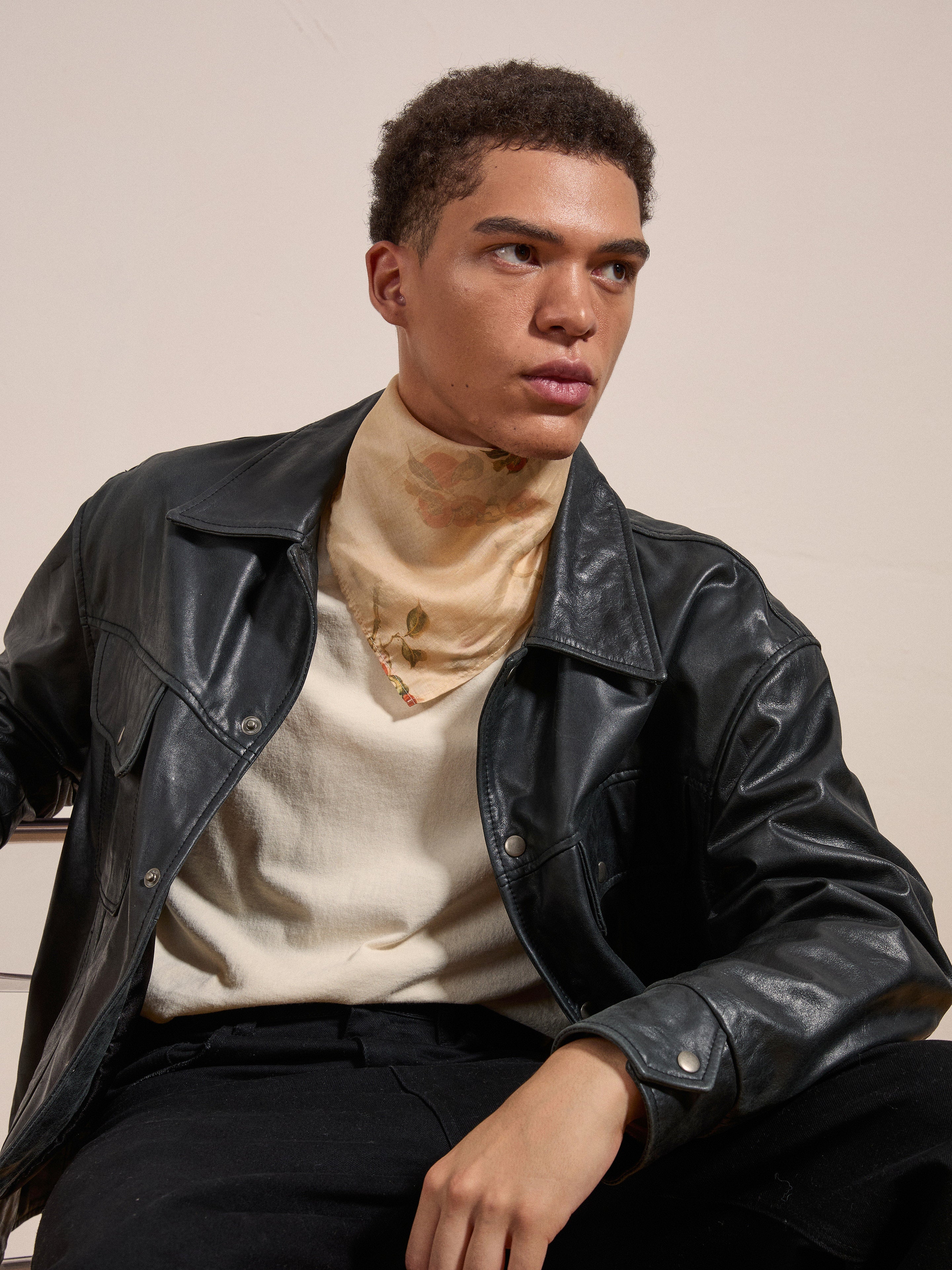 A person with short curly hair is seated, wearing a black leather jacket, a Roshan Floral Bandana by FOUND, a cream-colored shirt, and dark pants. They are looking off to the side against a plain background.