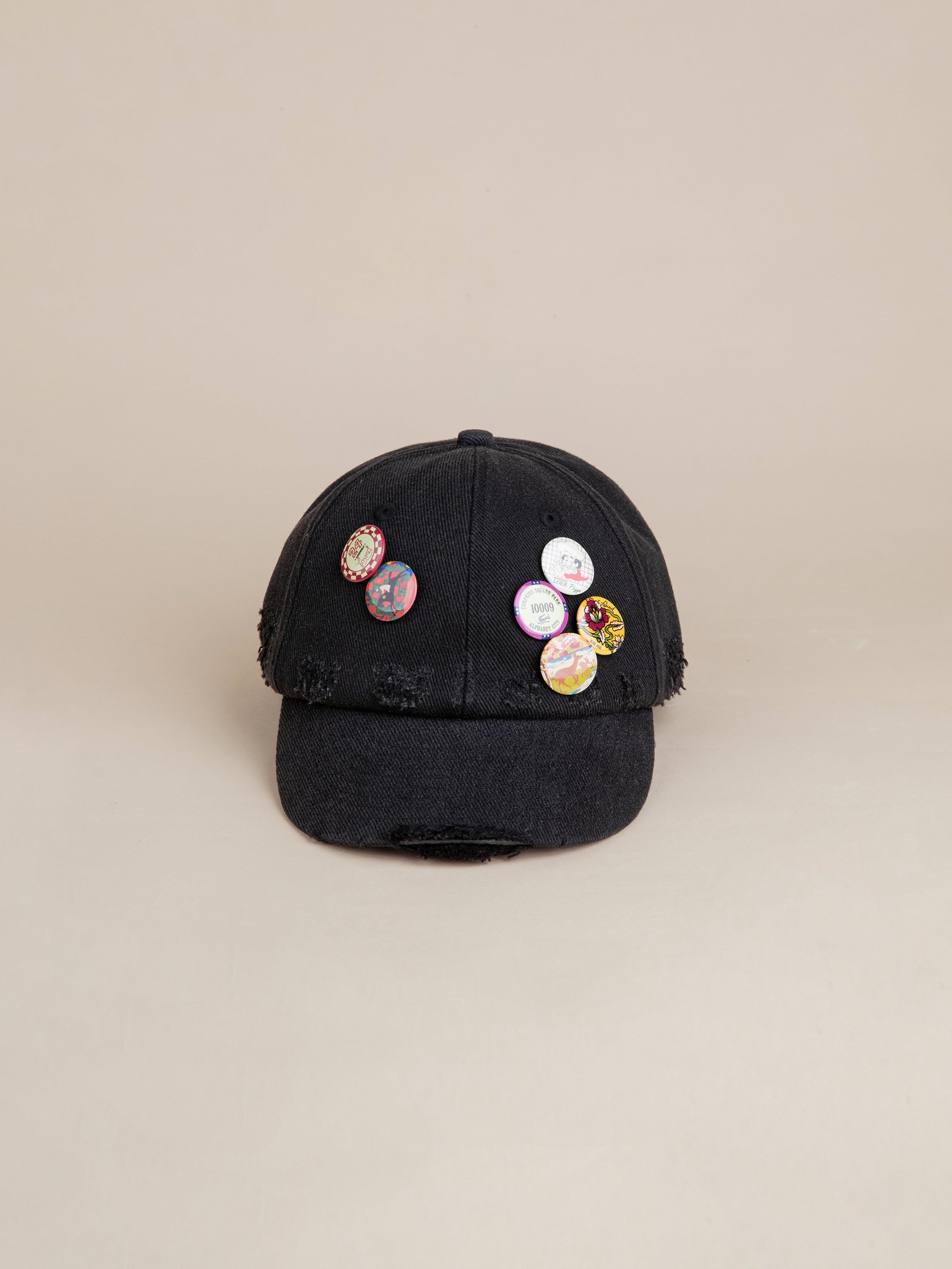 The FOUND Distressed Pin Cap, a black cotton hat featuring frayed edges and adorned with five vintage-inspired pins on the front, is displayed against a plain background.