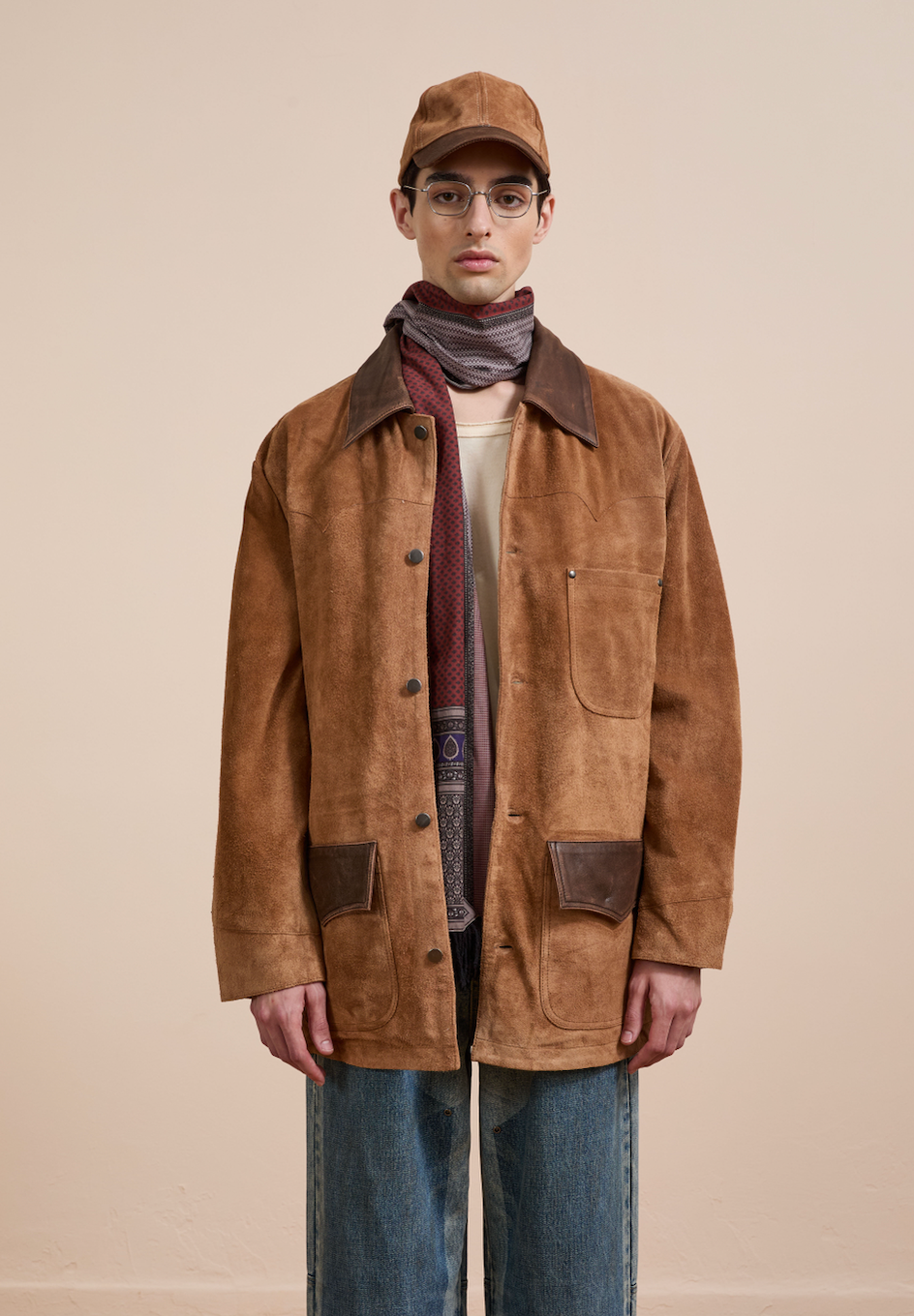 A person wearing the FOUND Suede Contrast Chore Jacket, a brown cap, glasses, scarf, and blue jeans stands against a plain beige background.