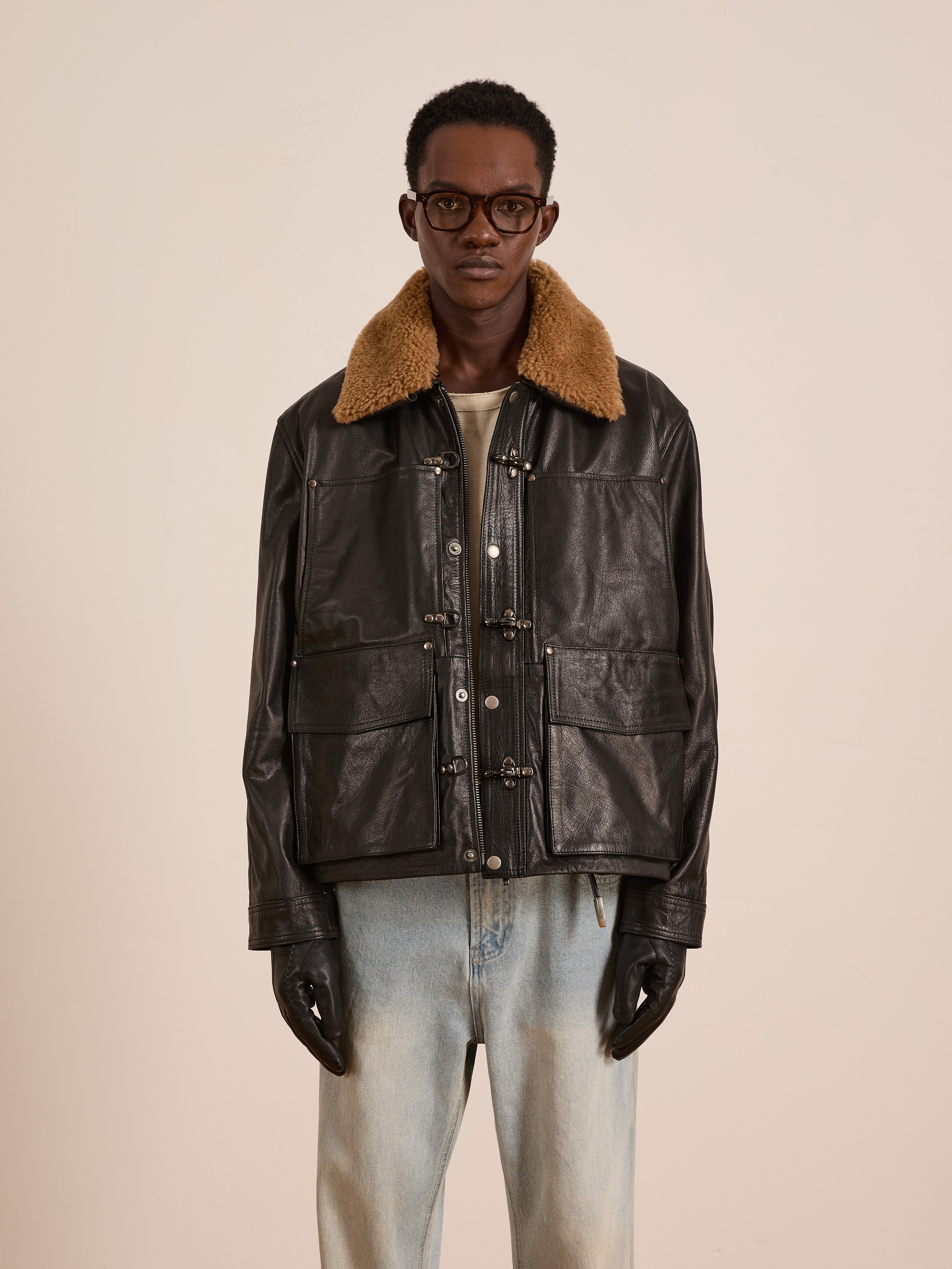 A person wearing glasses, black gloves, light blue jeans, and the FOUND Sherpa Collar Leather Utility Jacket stands against a plain light background.